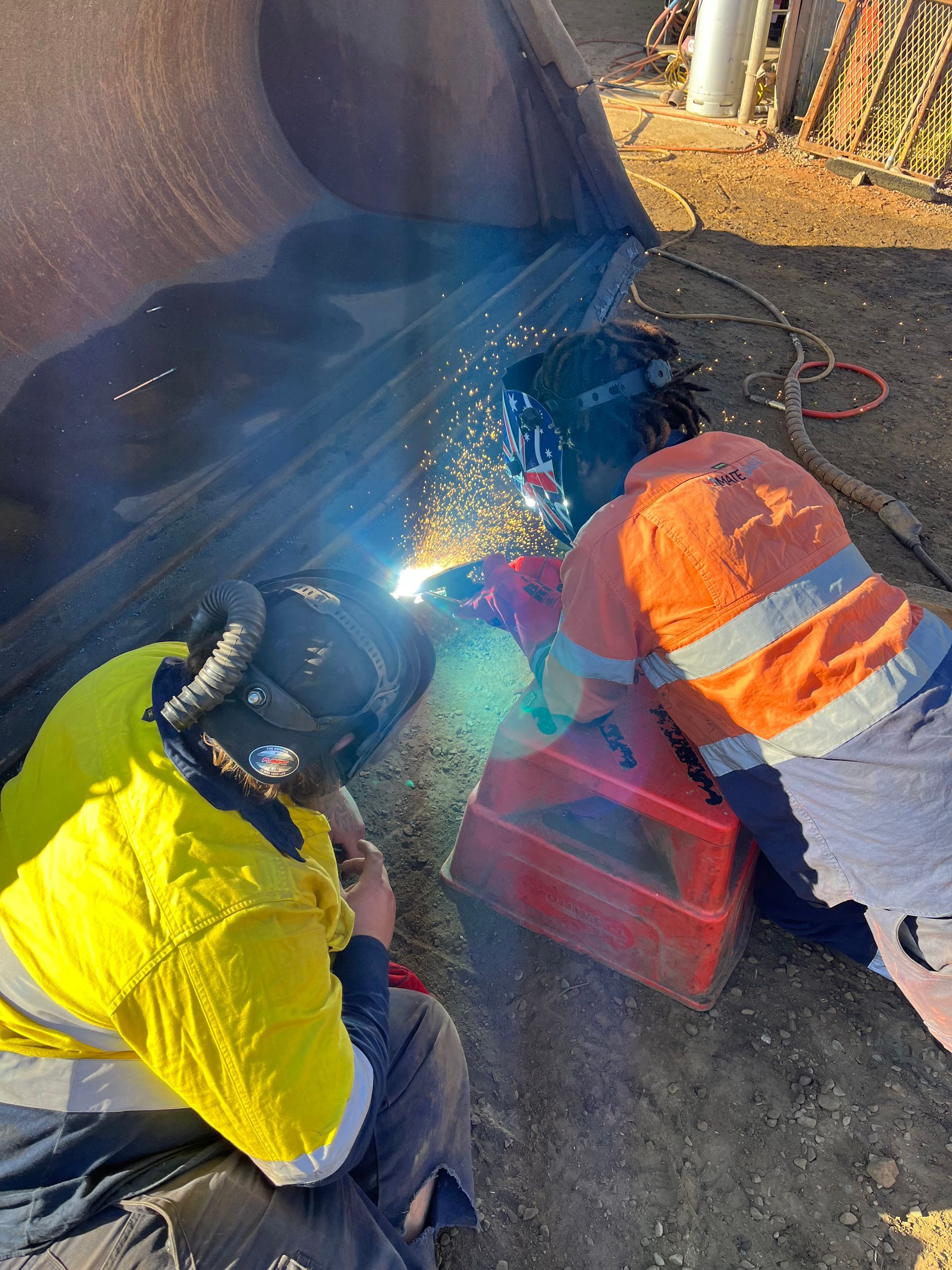Two people welding on a large metal pipe outdoors; one in yellow, one in orange safety gear.