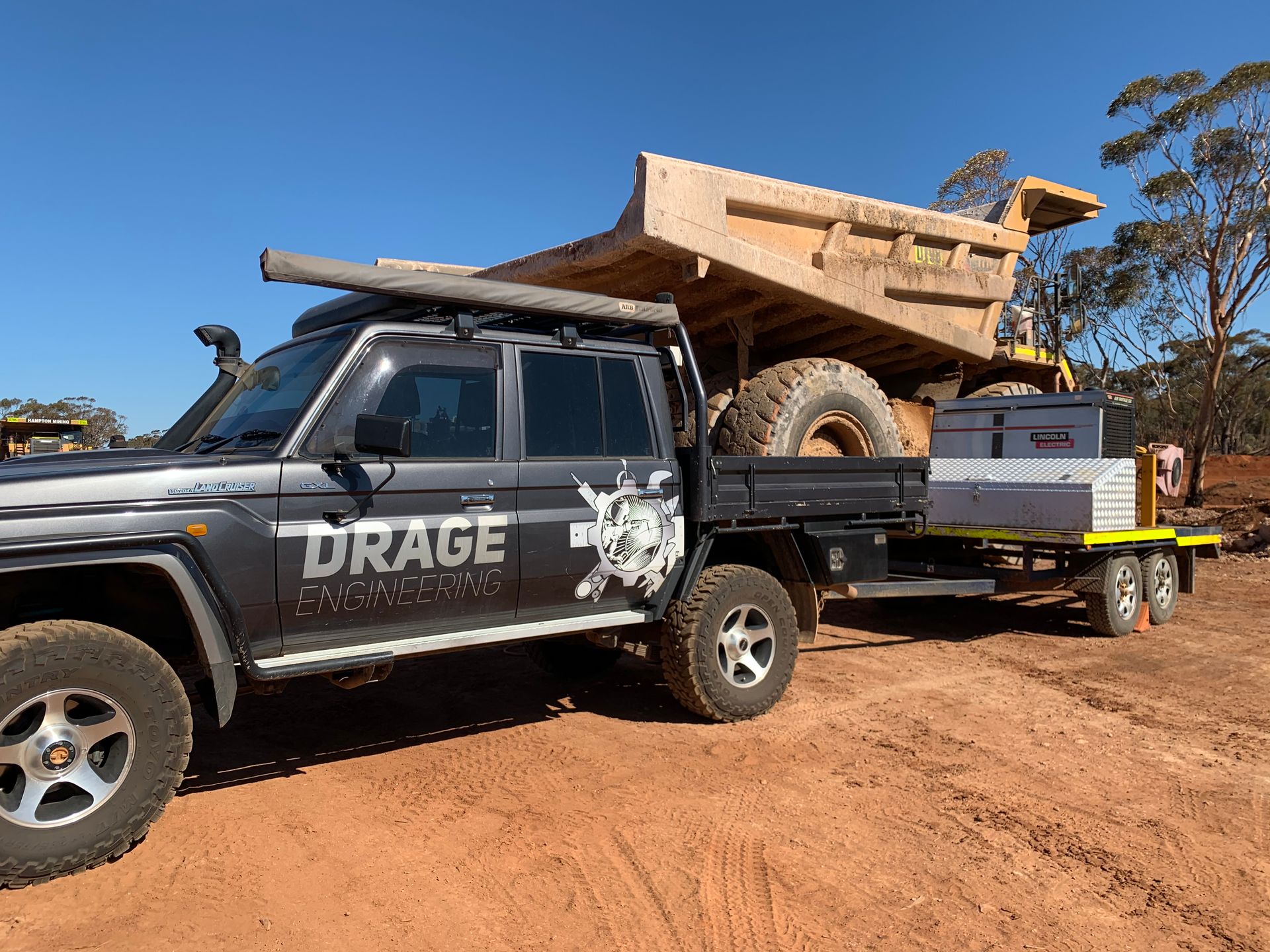 Gray truck towing trailer with large earthmoving truck tire in a dirt field.