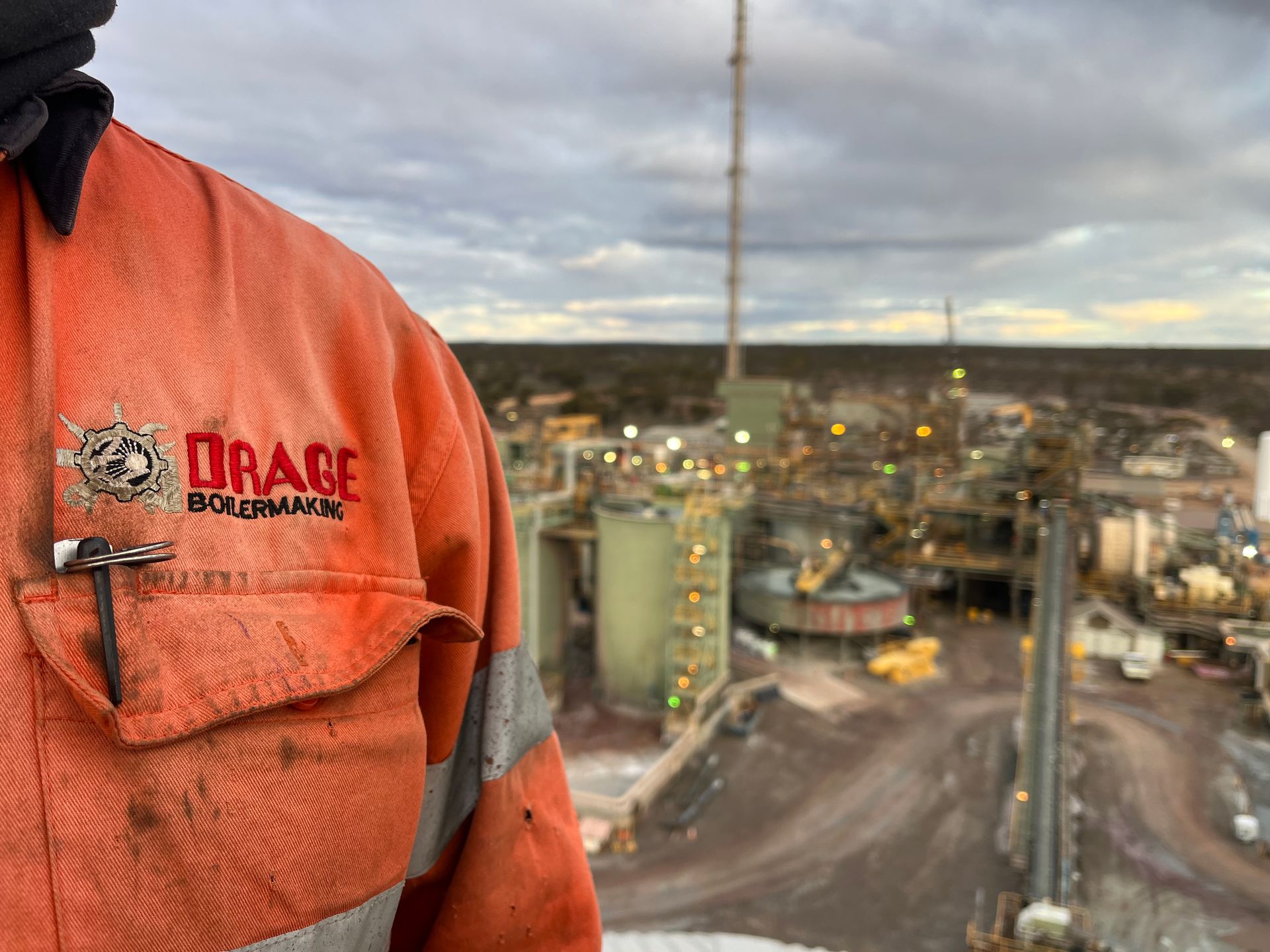 Orange work jacket with logo, overlooking industrial facility at dusk.