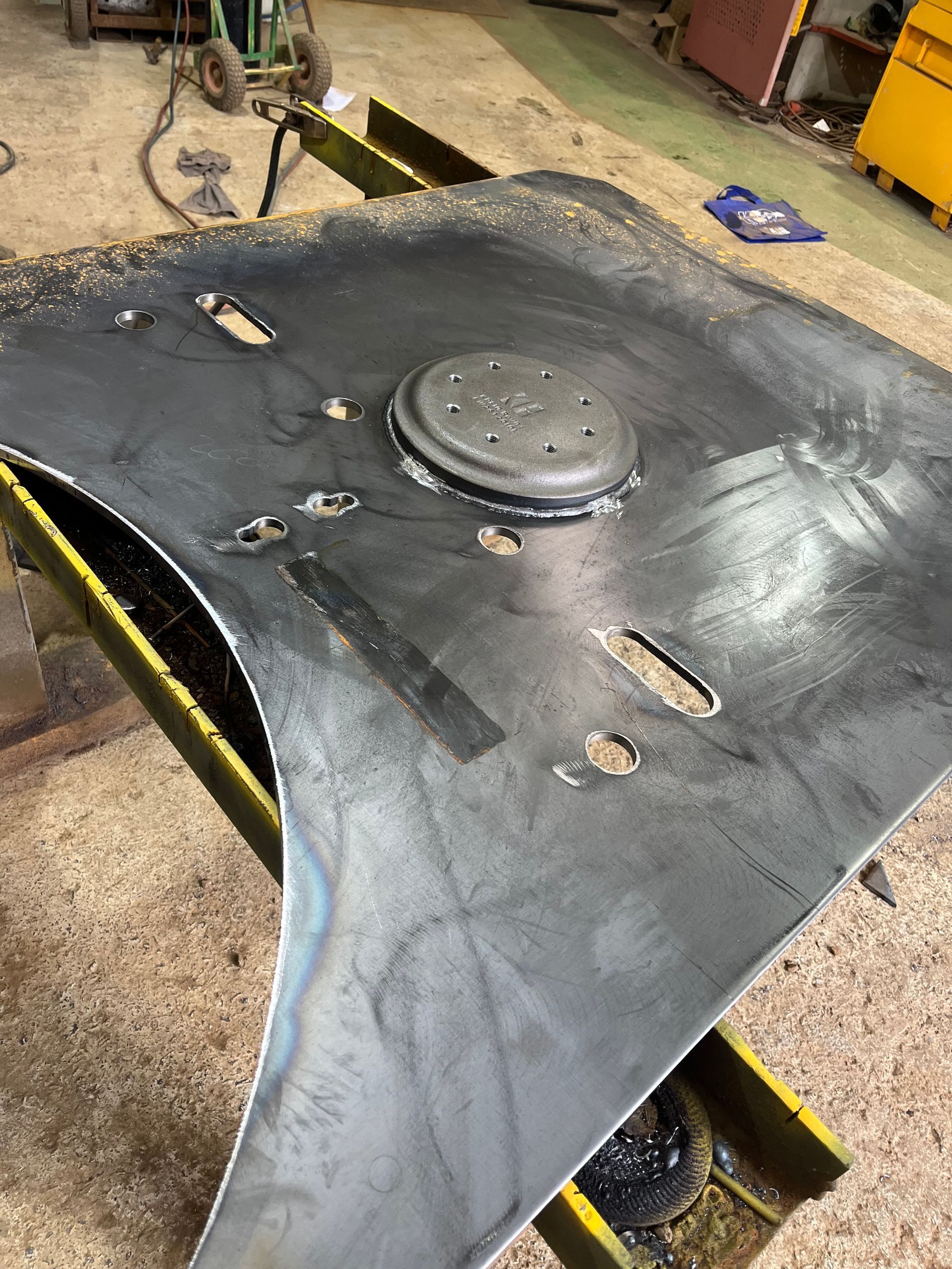 Black metal panel with welded circular feature and cut-out slots, resting on yellow support beams.