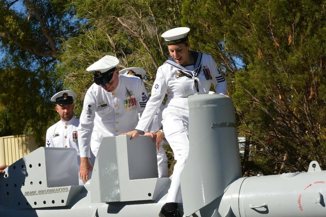 Sailors in white uniforms, helping each other on a gray structure outdoors under sunlight.