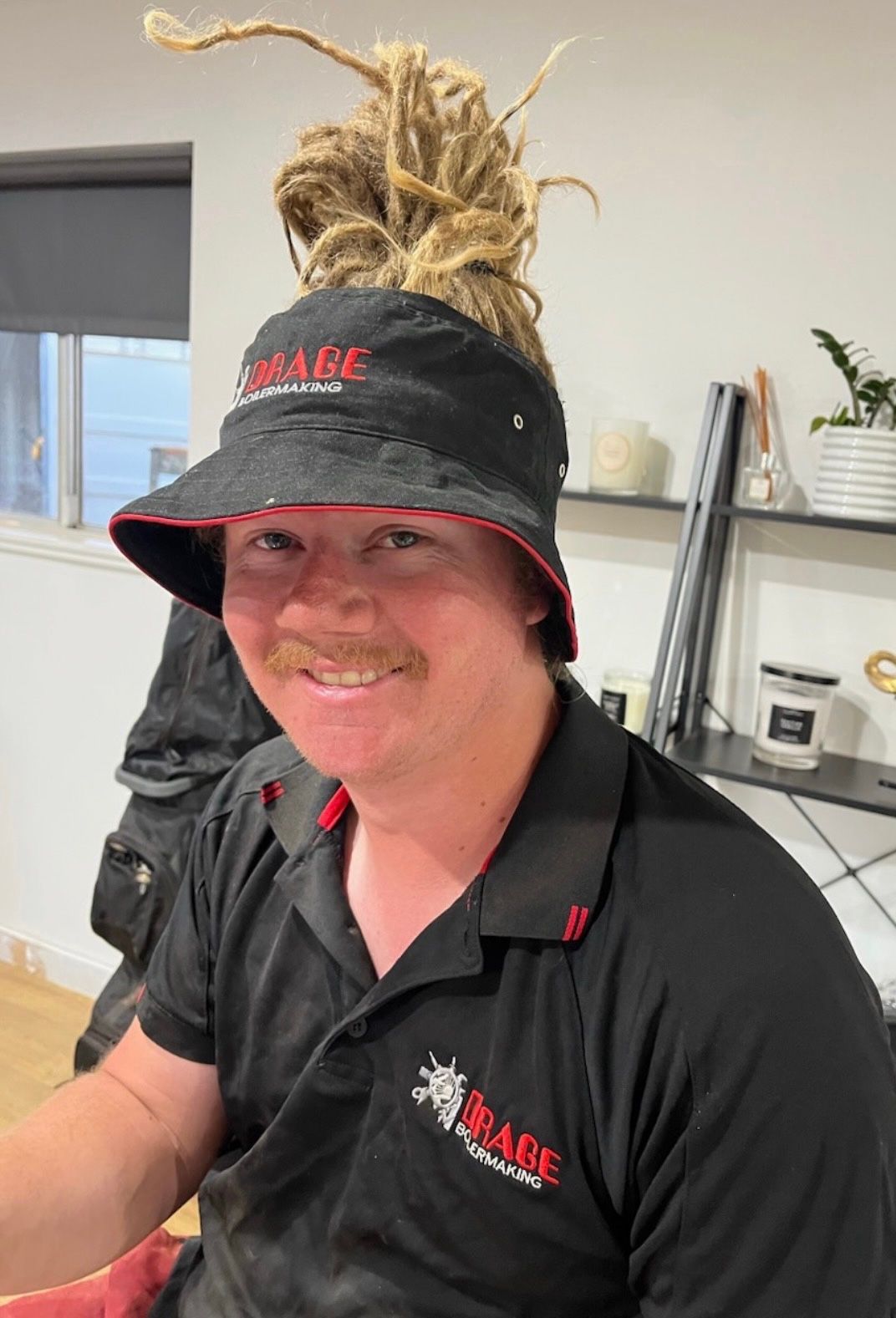 A smiling person wearing a black visor with dreadlocks piled on top and a branded black polo shirt in an indoor setting.