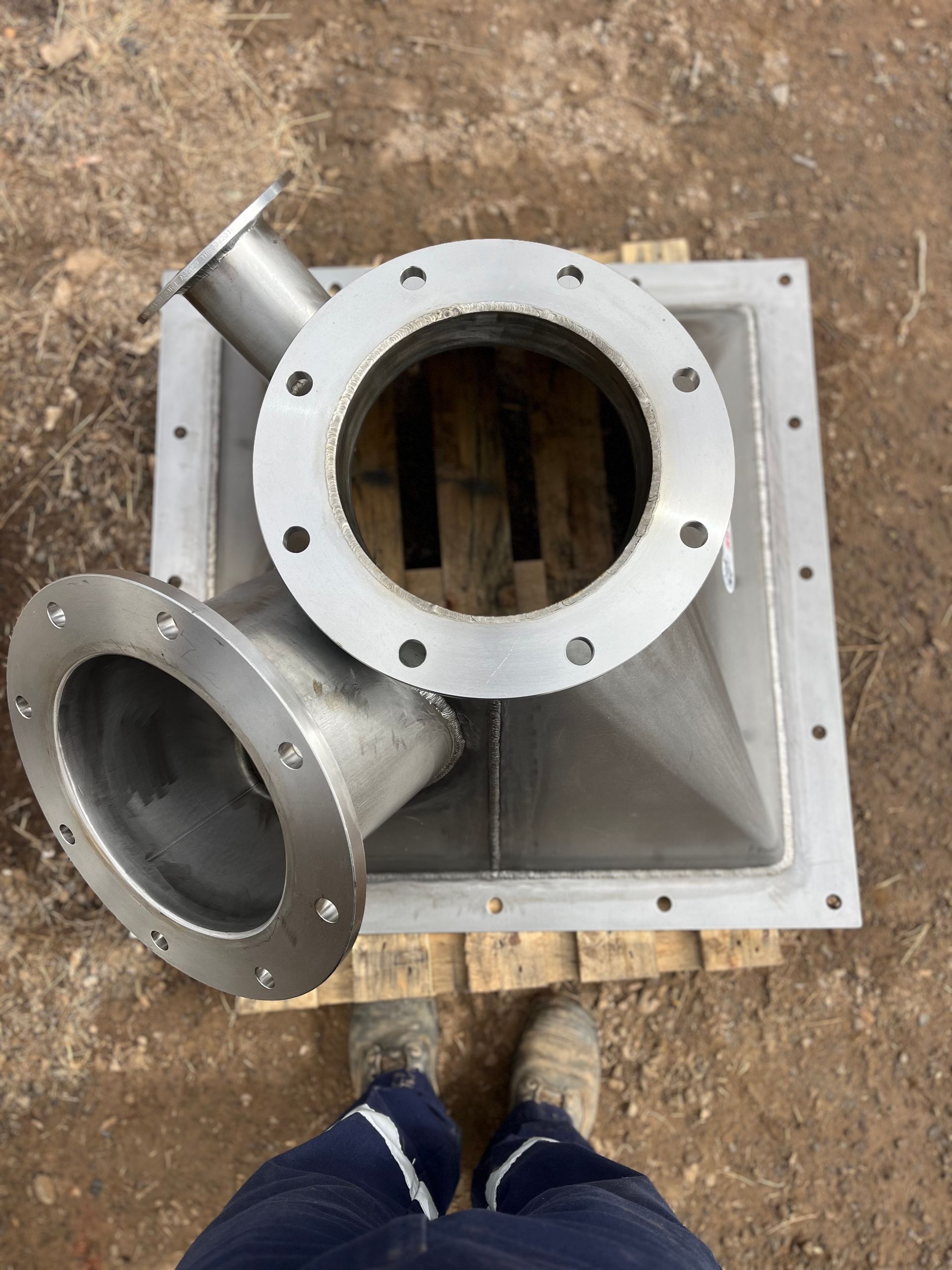 Stainless steel industrial part with flanges, sitting on a wooden pallet; a person's feet are visible.