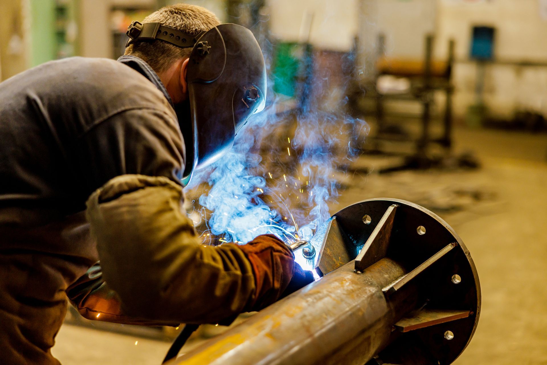 Welder in protective gear welding metal, creating sparks in an indoor industrial setting.