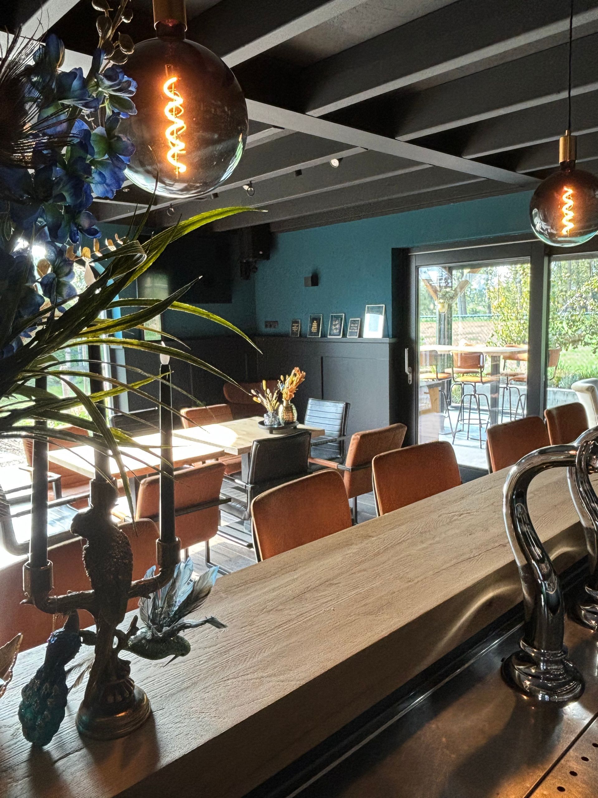 Gezellig café-interieur met hangende pendellampen, houten tafels en grote ramen die daglicht binnenlaten.