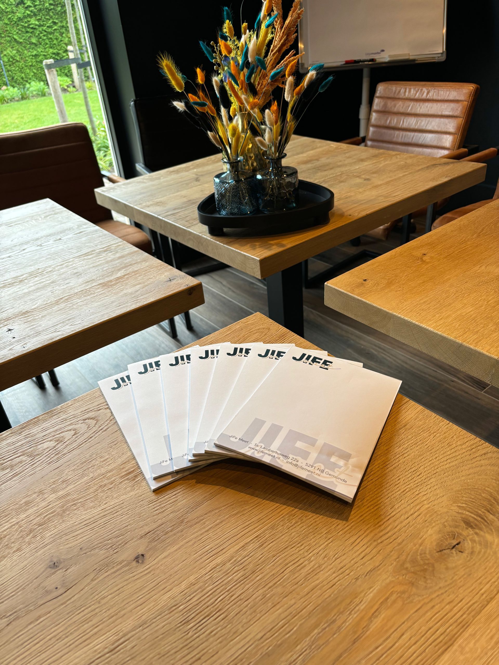 Visitekaartjes met logo liggen uitgespreid op een houten tafel in een moderne kamer met een tafeldecoratie en stoelen.