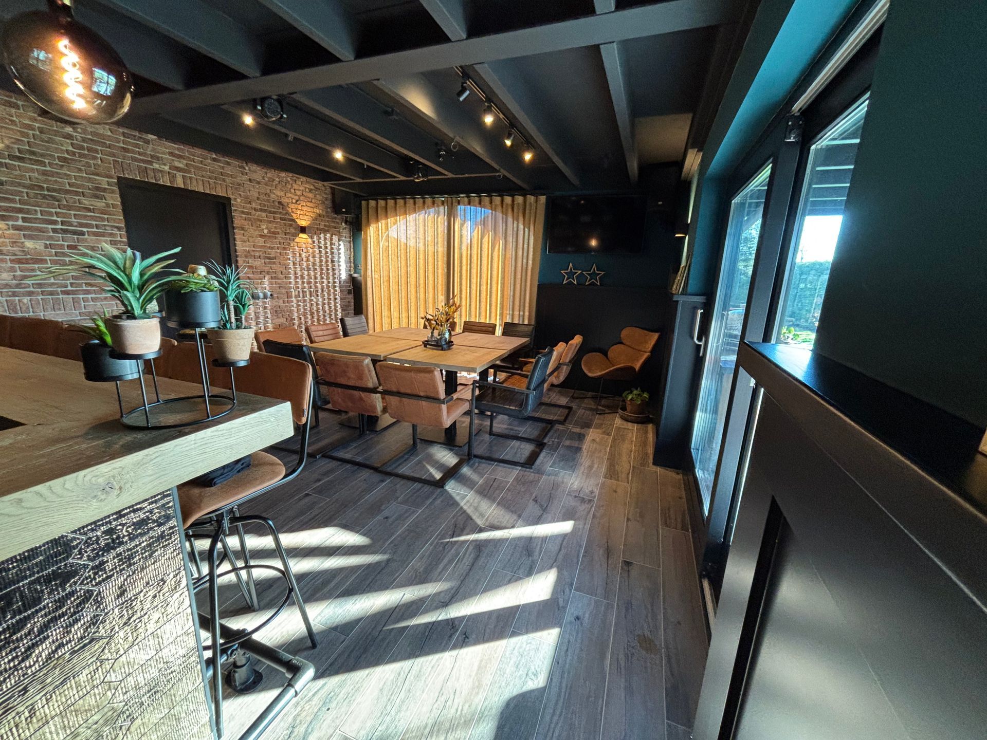 Modern interieur met houten tafels, zwarte stoelen, bakstenen muren en zonlicht dat door grote ramen naar binnen valt.