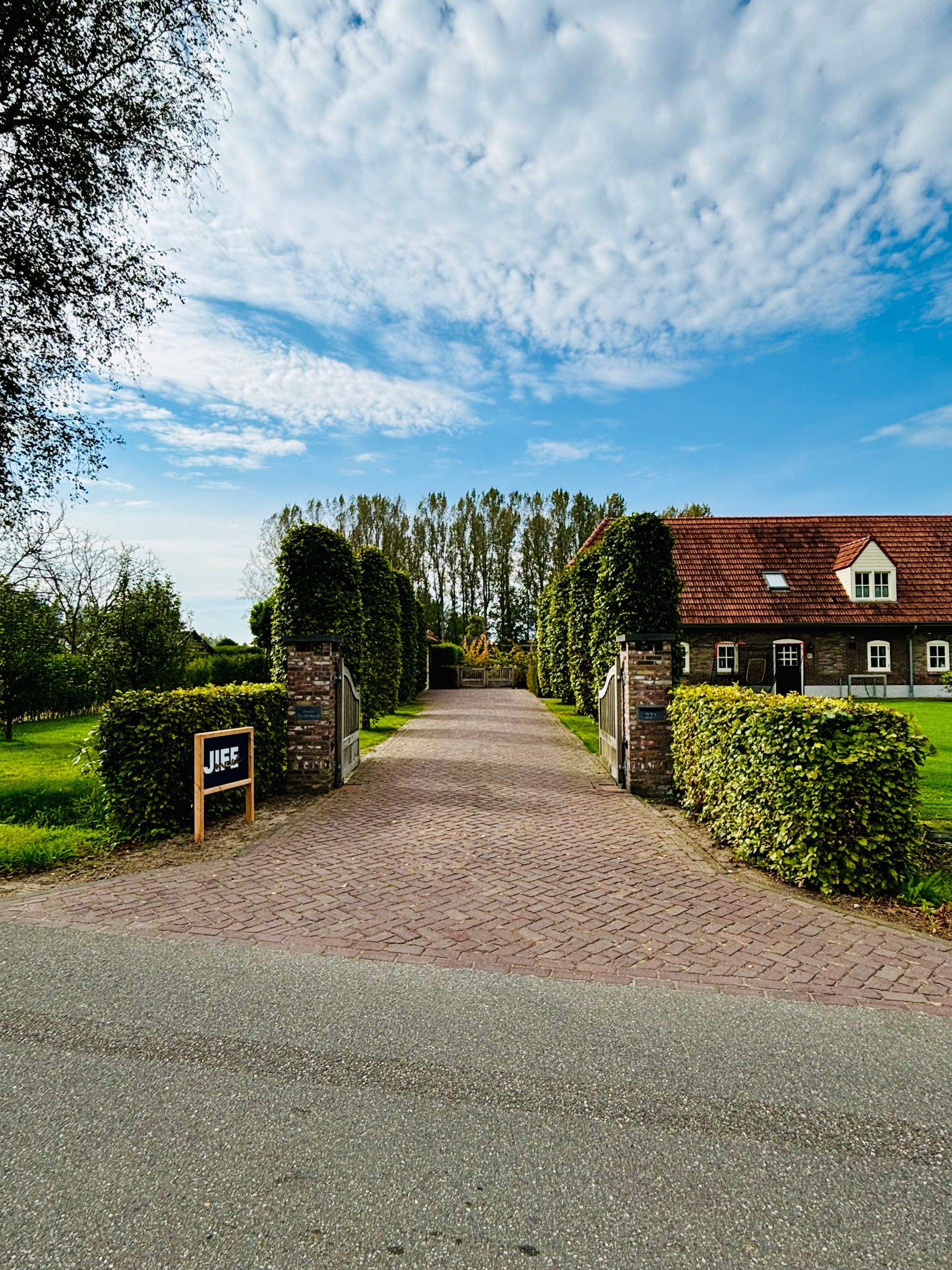 Een bakstenen oprit leidt naar een huis met een rood dak, omgeven door hagen en bomen, onder een gedeeltelijk bewolkte hemel.