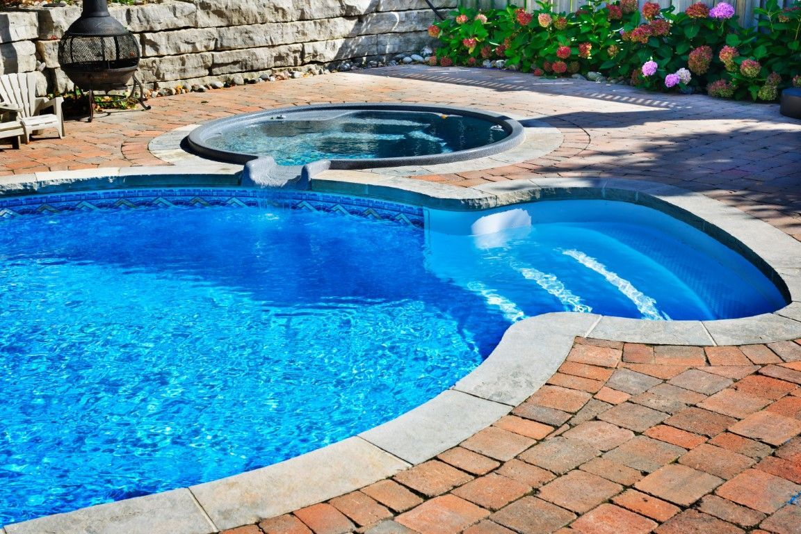Backyard pool with bright blue water, hot tub, stone wall, and brick patio beside flowers