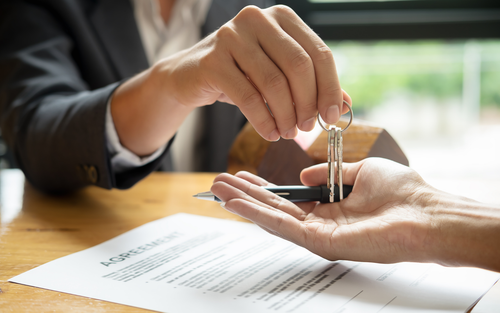 A person in a suit hands keys to another person over a signed agreement on a wooden table.