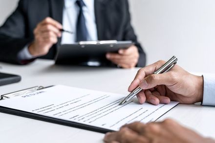 A person writing on a document while sitting across from an interviewer holding a clipboard at a desk.