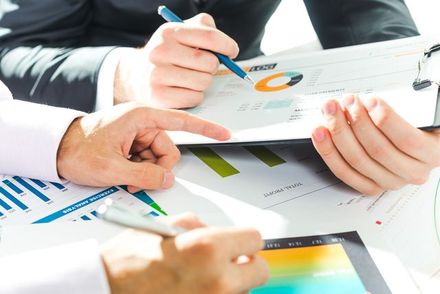 Business professionals collaborate over financial charts, graphs, and tablets in a well-lit office setting.