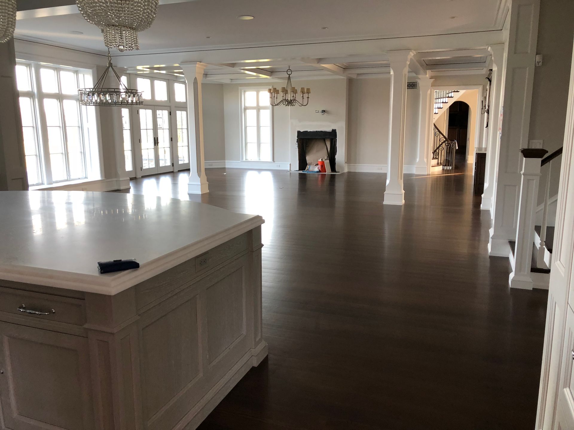 A spacious open-plan room with a large kitchen island in the foreground, white columns, dark wood floors, and a fireplace.