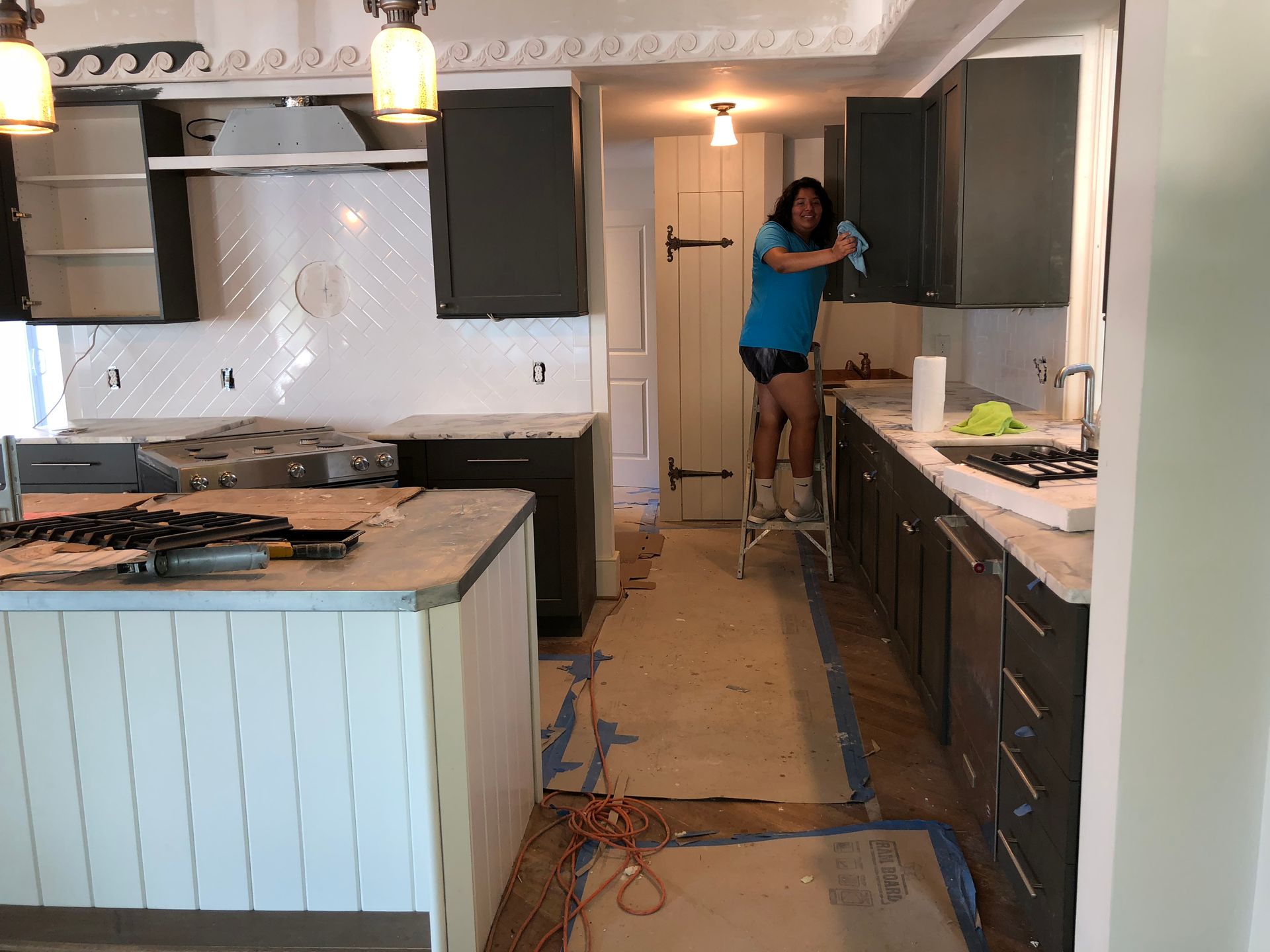Woman cleaning kitchen window with yellow gloves. Wooden blinds and countertop in bright kitchen.