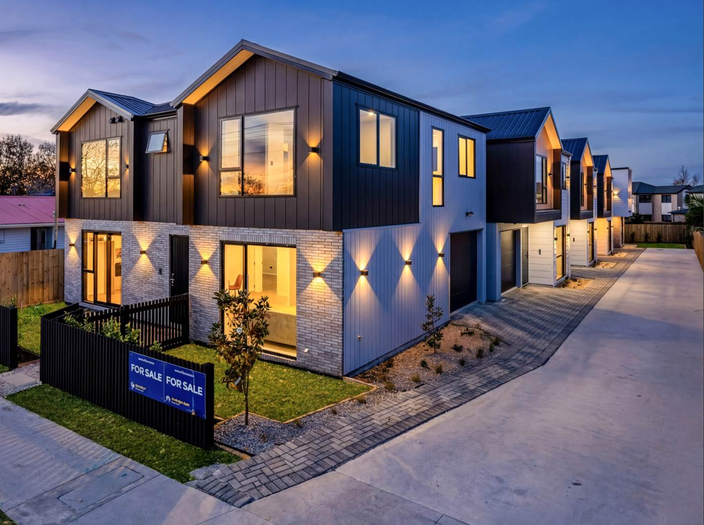 Modern two-story apartment building at dusk with lit windows, dark siding, and a sidewalk driveway