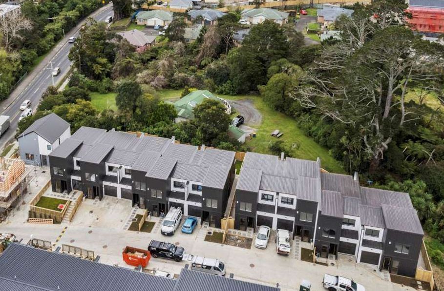 Aerial view of modern dark gray townhouses beside a road, with parked cars and green trees behind.