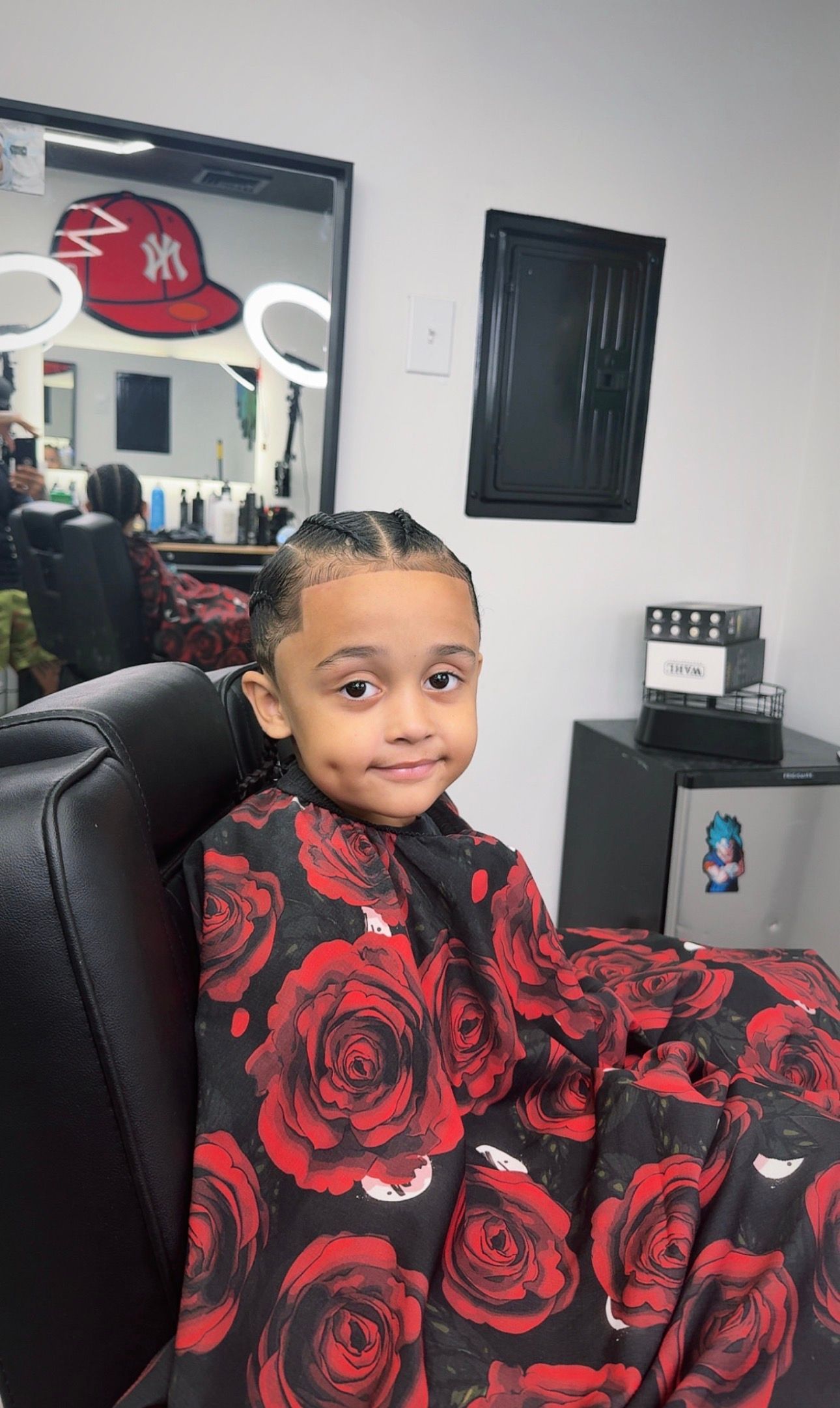 Child with braided hair smiles, wearing a rose-patterned barber cape, seated in a barber chair.