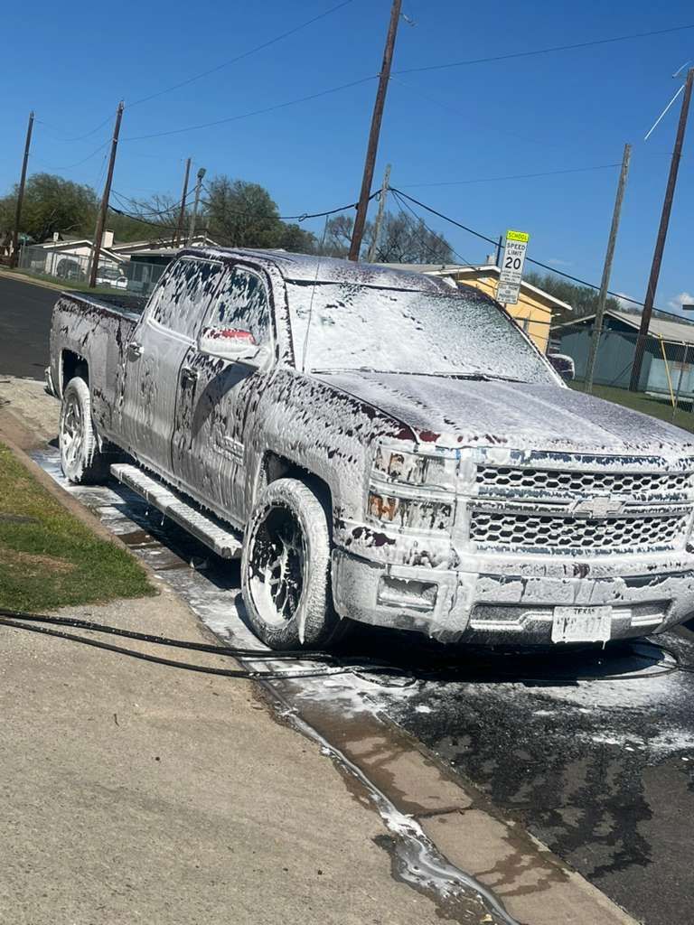 Foam-covered silver pickup truck parked on a sidewalk beside a road, being washed outdoors.