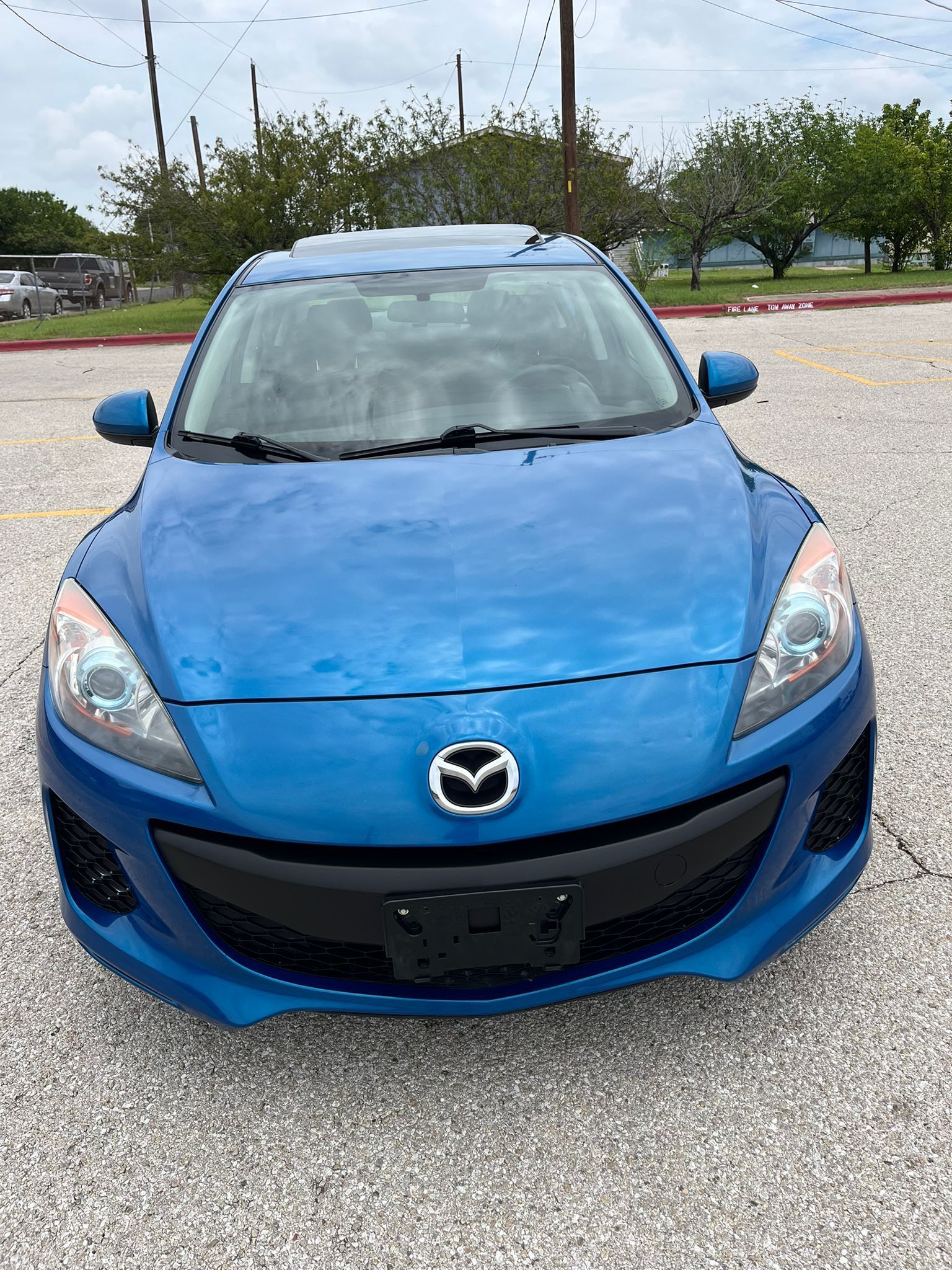 Front view of a blue Mazda hatchback parked on a gravel lot under cloudy skies