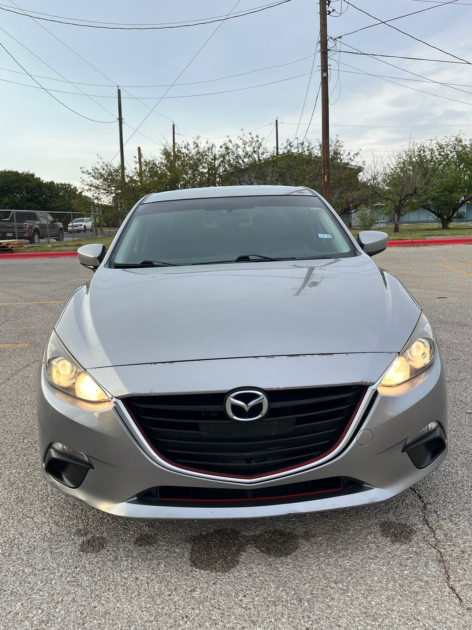 Front view of a silver Mazda sedan parked outdoors at dusk with headlights on