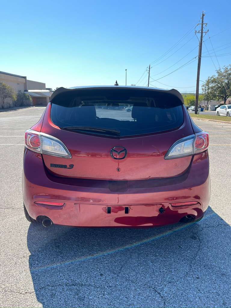 Rear view of a red hatchback with bumper damage parked on a sunny roadside.