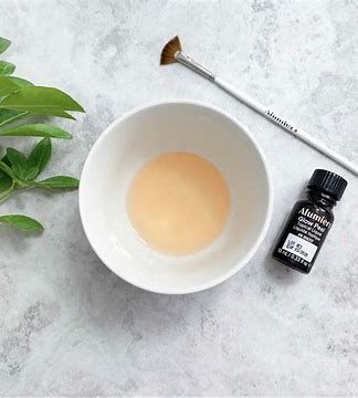 White bowl with beige liquid, makeup brush, and small black bottle on a light gray surface