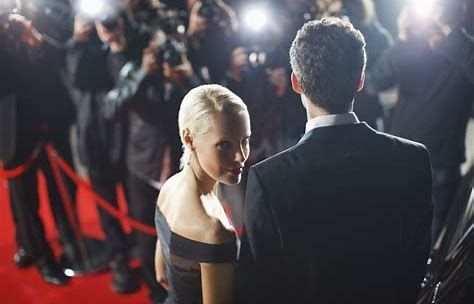Woman in an off-shoulder black dress looking back at a man on a red carpet with photographers flashing behind them