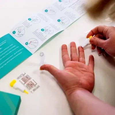 Hand using a lancet to prick a finger for a blood glucose test on a white table with test kit instructions.