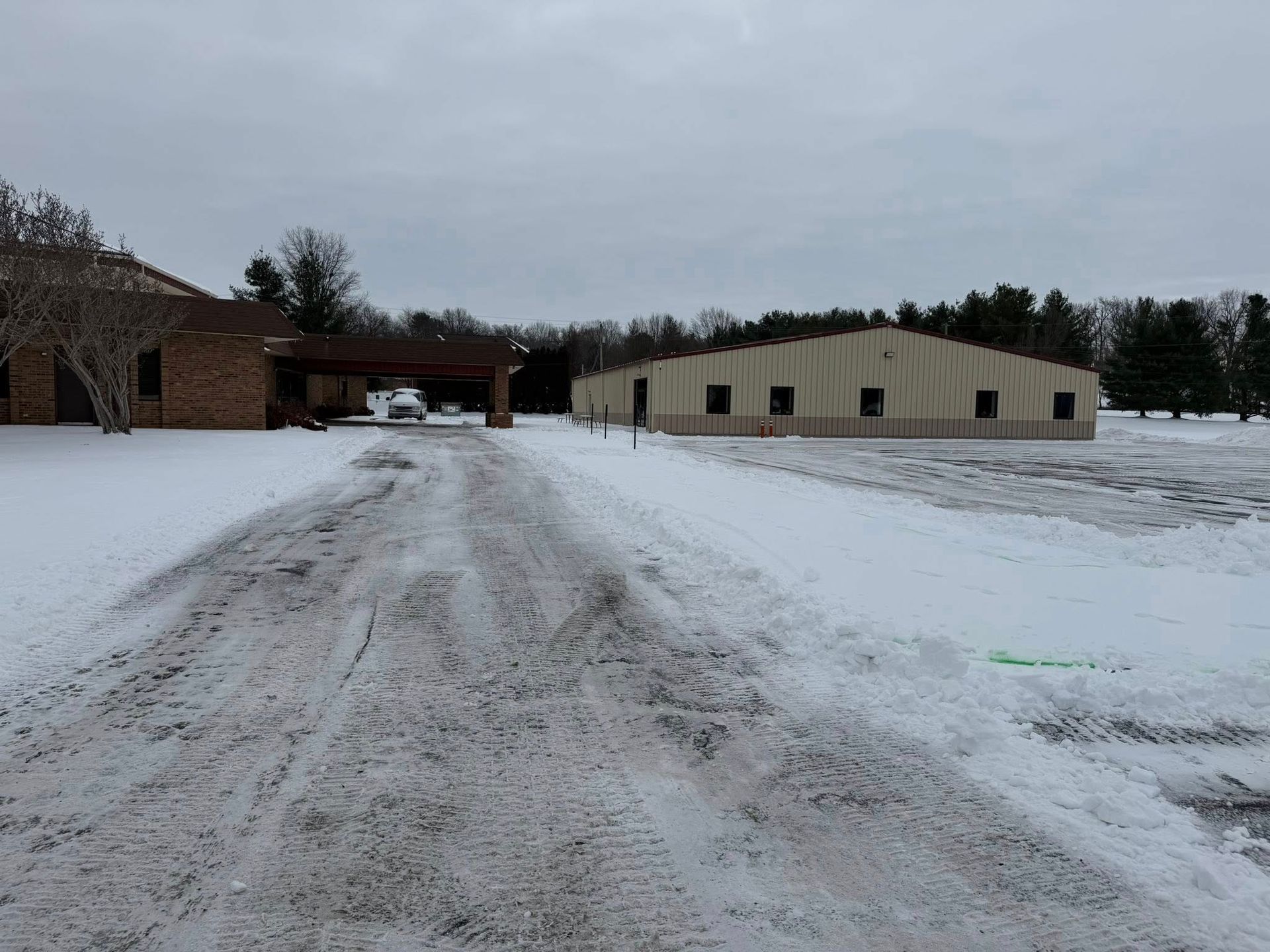 Snowy driveway leading to a farmyard with barns and a beige building under a gray sky