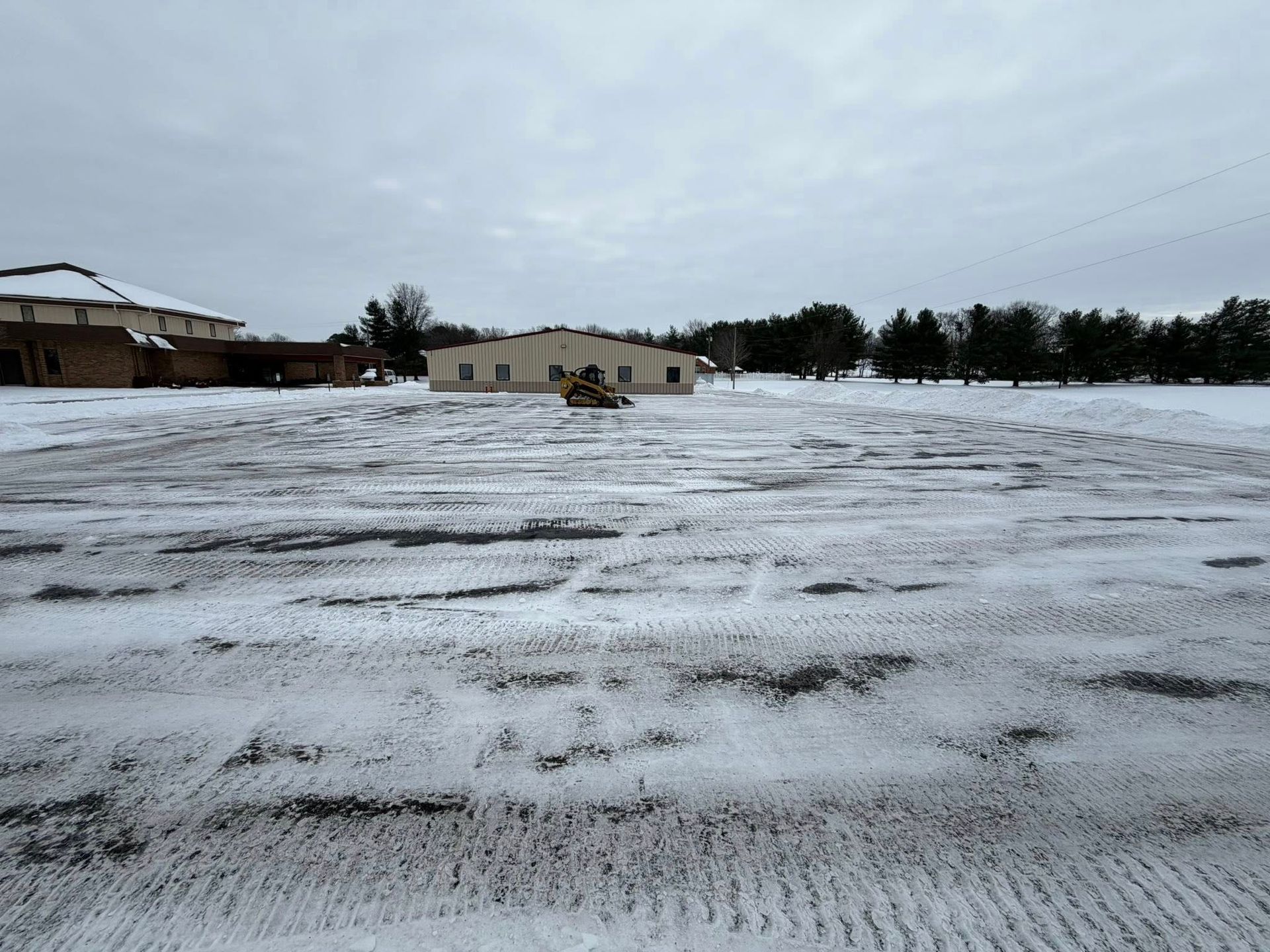 Snow-covered open field with a few buildings and trees under a cloudy sky