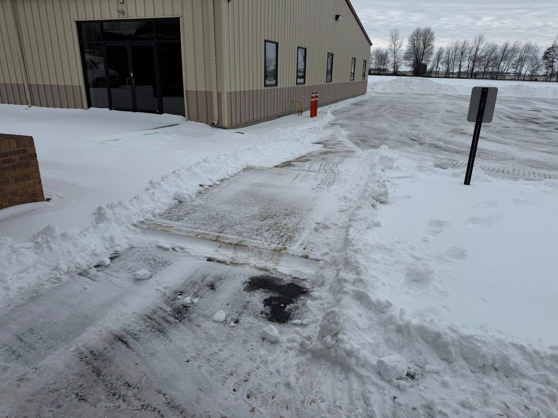 Snowy walkway beside a beige building with cleared pavement and an orange fire extinguisher near the entrance