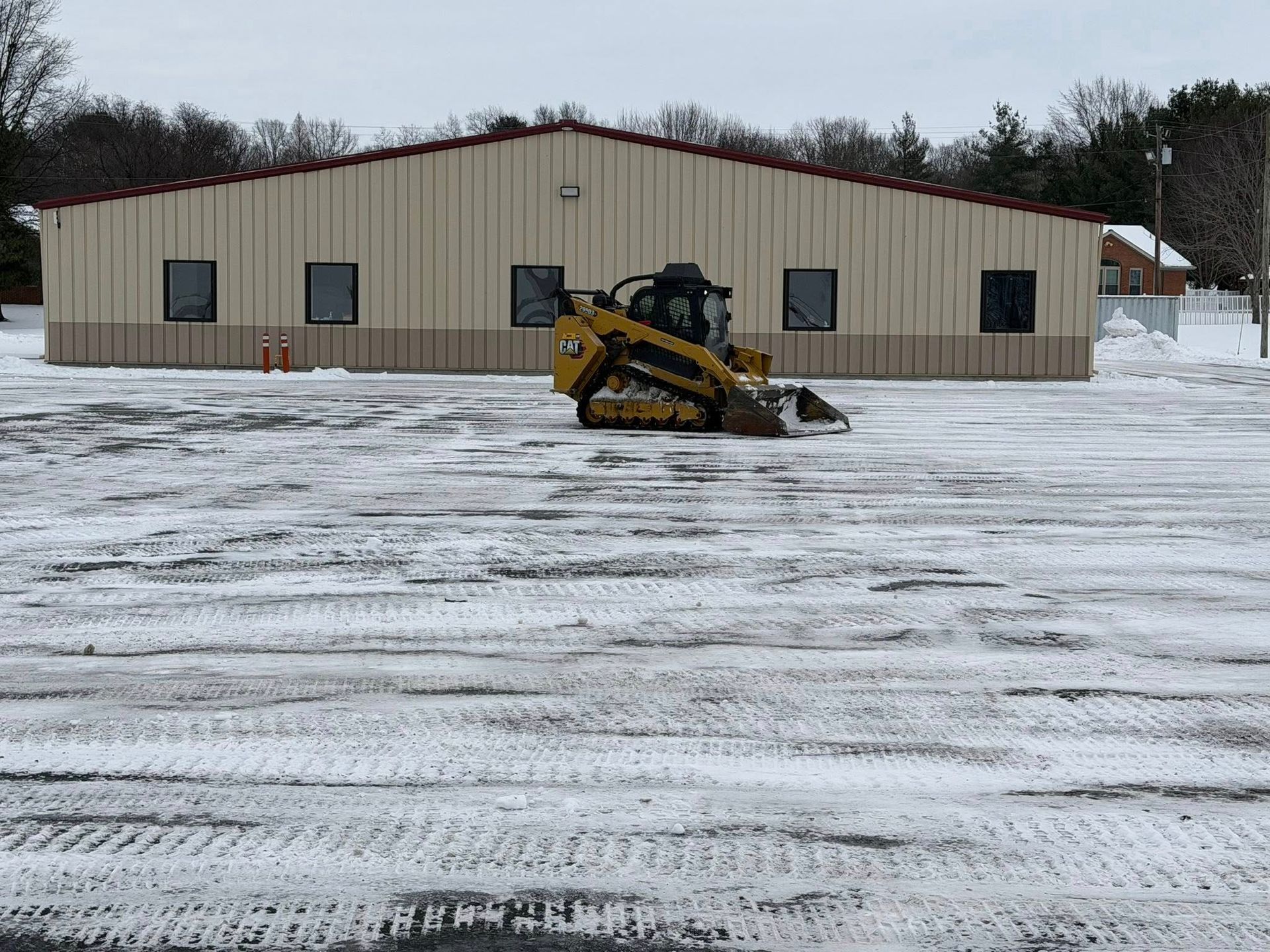 Yellow skid steer plowing snow in front of a beige metal building on a snowy lot.