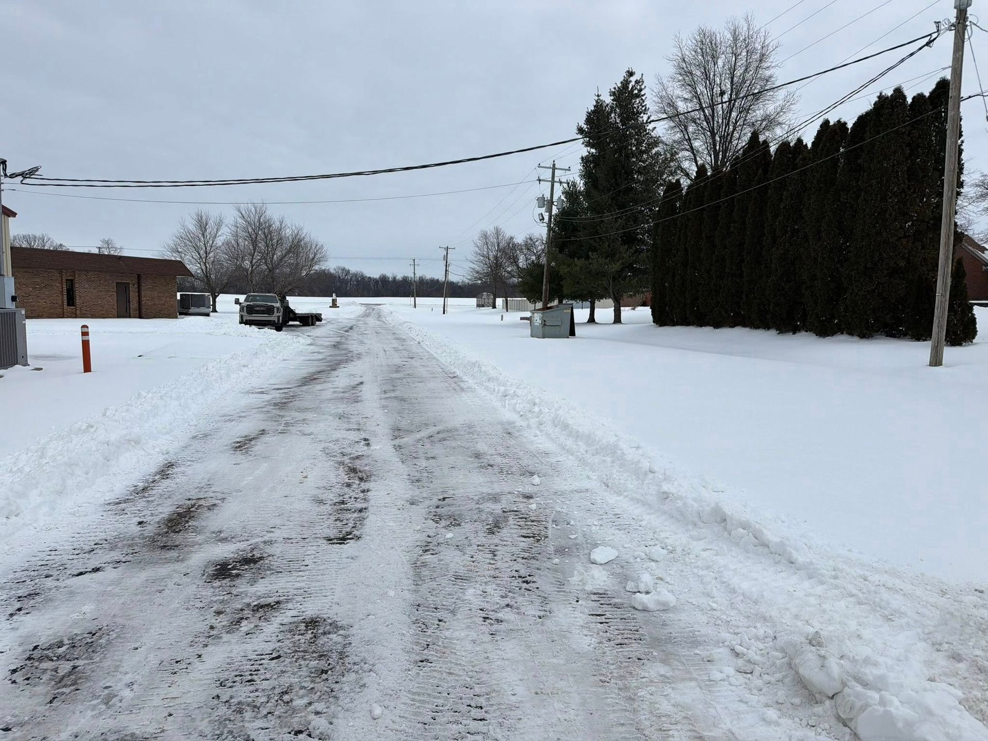 Snow-covered rural road with tire tracks, lined by trees and a brick building under a gray sky