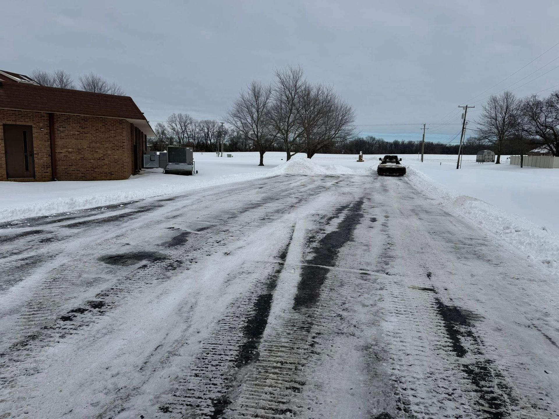 Snow-covered road with tire tracks leading past a brick building and bare trees under a gray sky.
