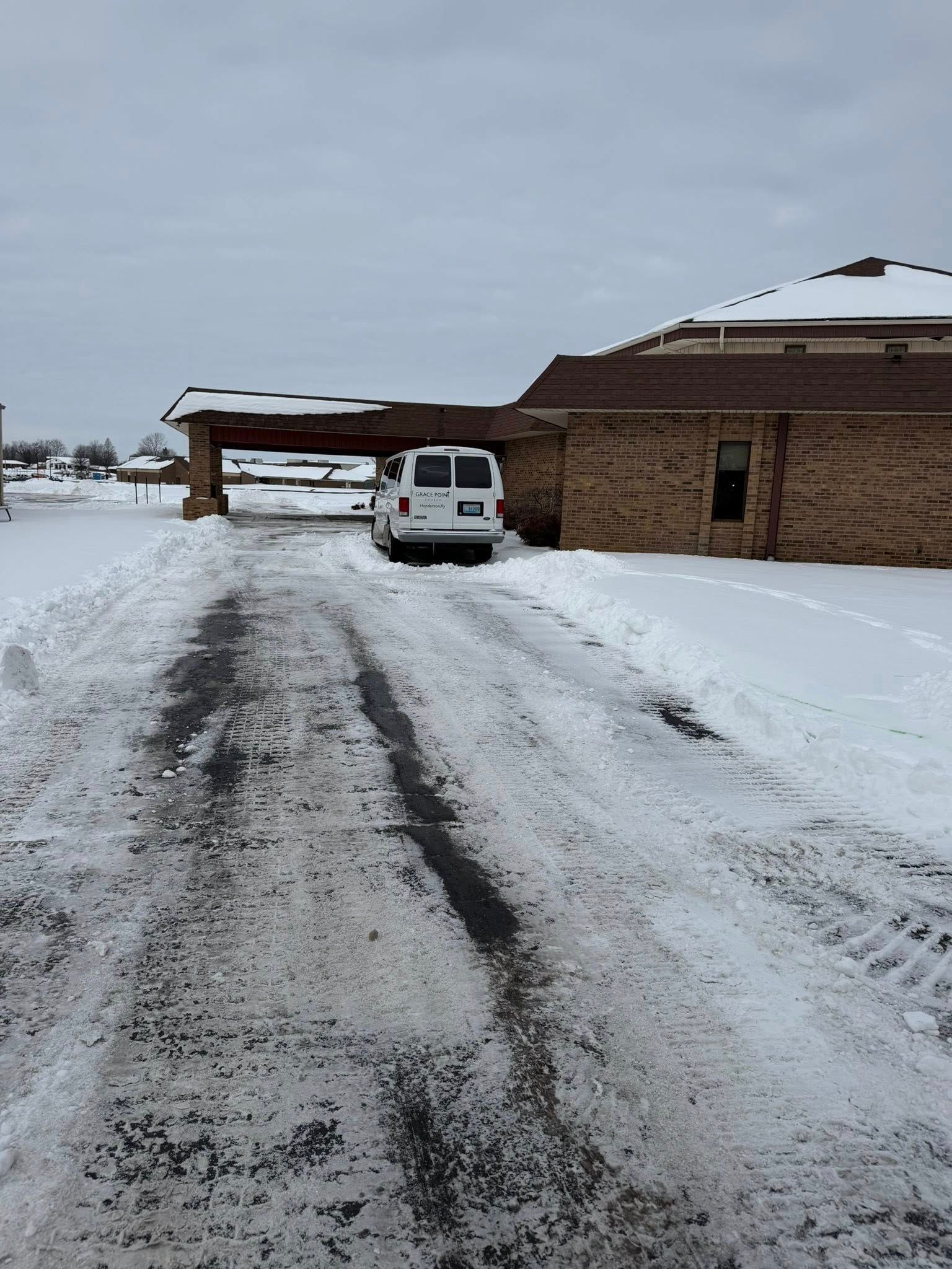 Snowy driveway with a white SUV parked beside a brick house under an overcast sky