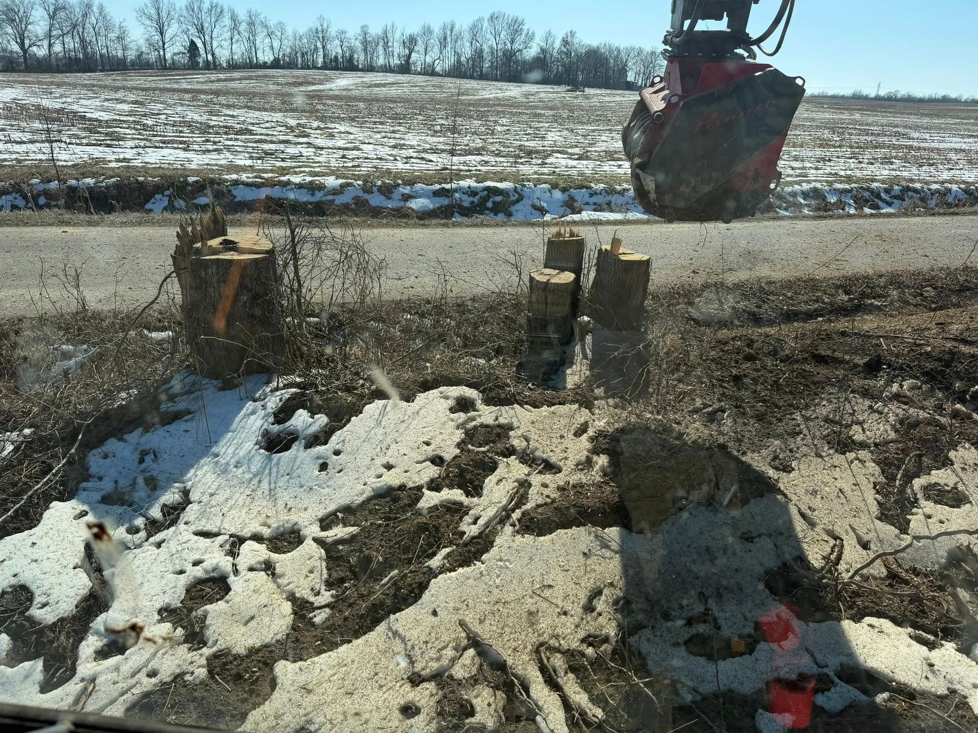Excavator bucket dumping soil onto a snowy roadside work area beside a trench