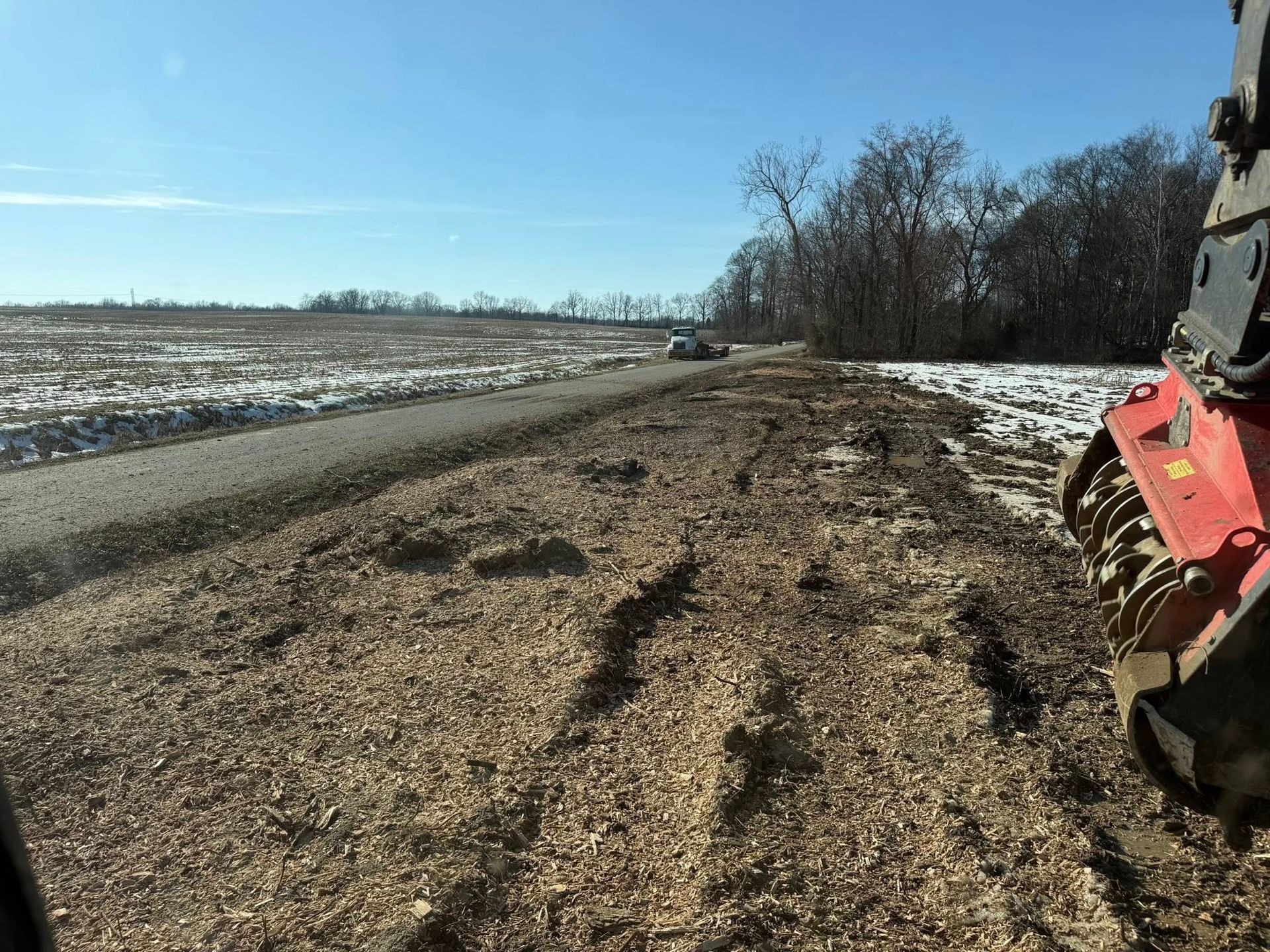 Rural dirt road beside snowy fields, with a red tractor at the right edge under a clear blue sky