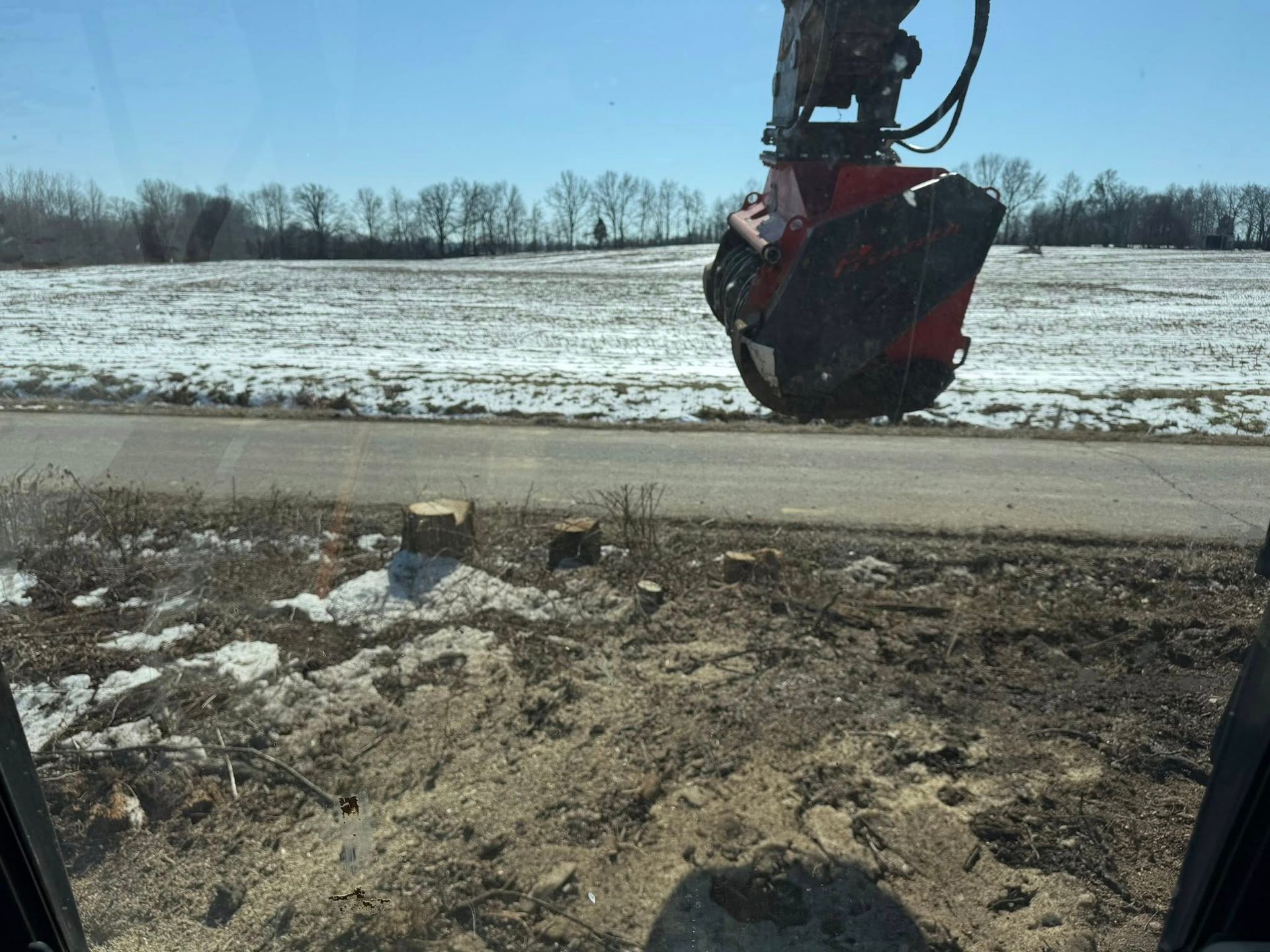 Excavator claw hanging over a snowy roadside beside a muddy field
