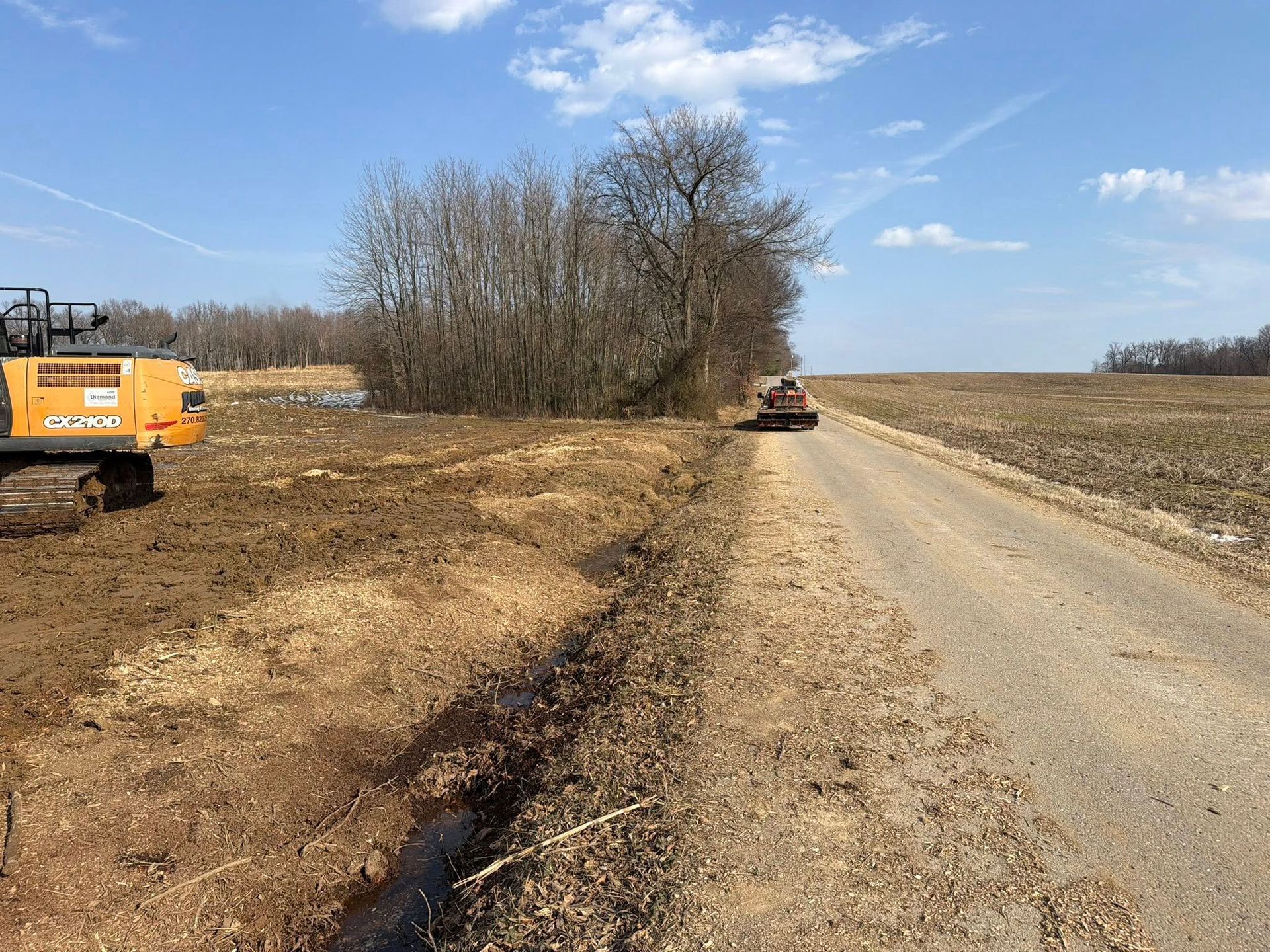 Rural dirt road with an excavator on the left and a tractor in the distance under a clear blue sky