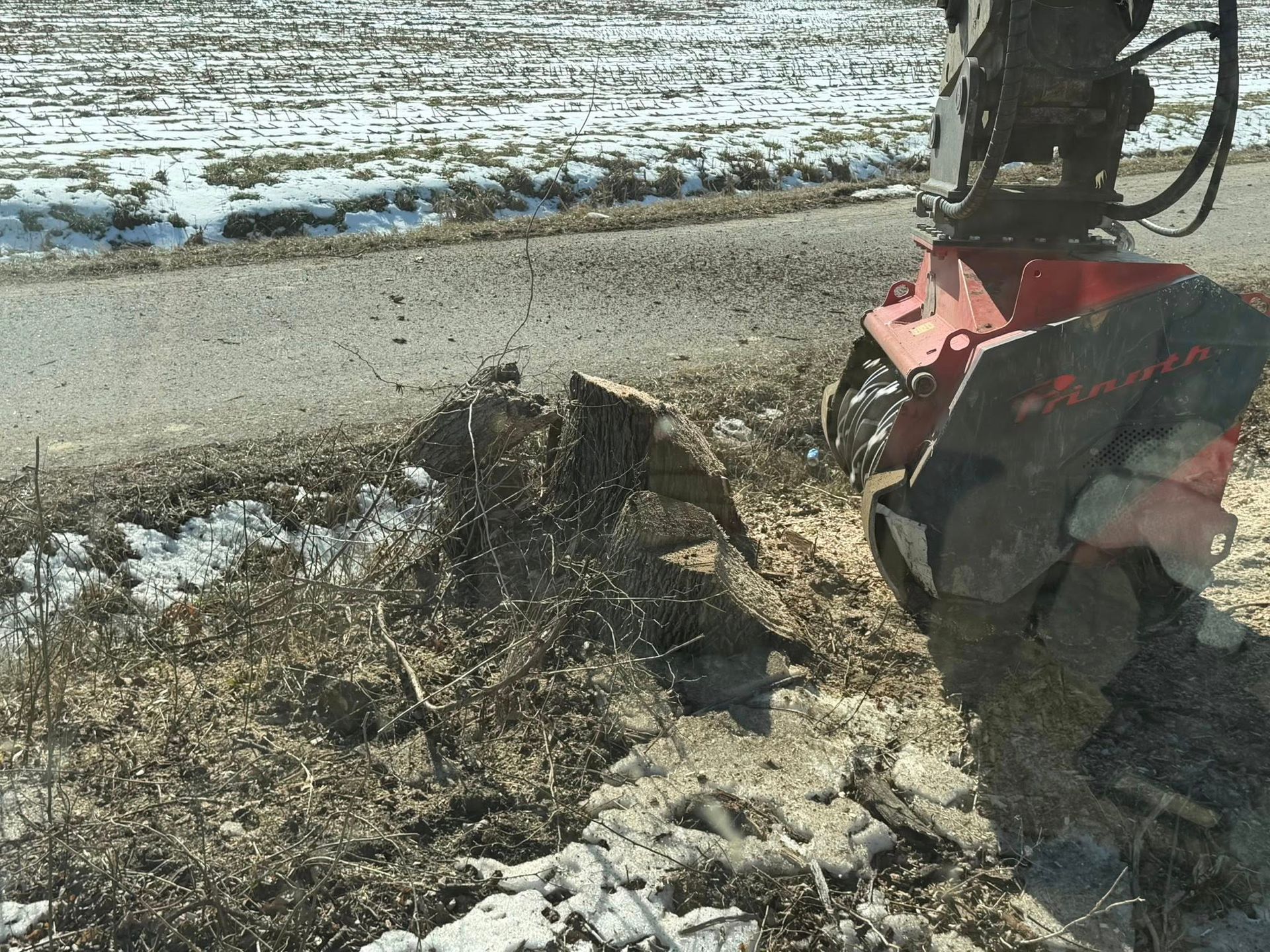 Red snow blower clearing a muddy roadside with snow patches