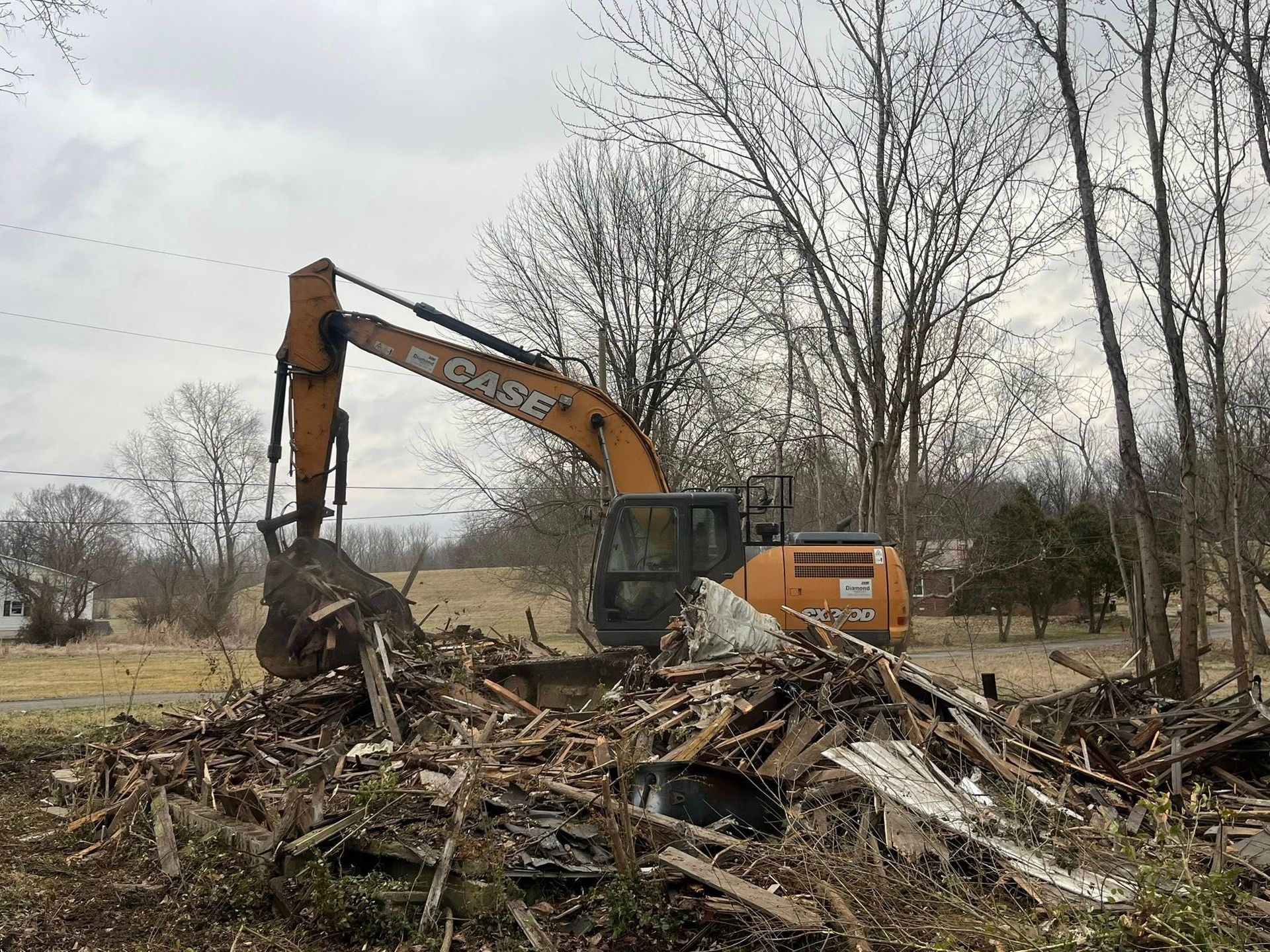 Orange excavator clearing debris in a leafless wooded lot