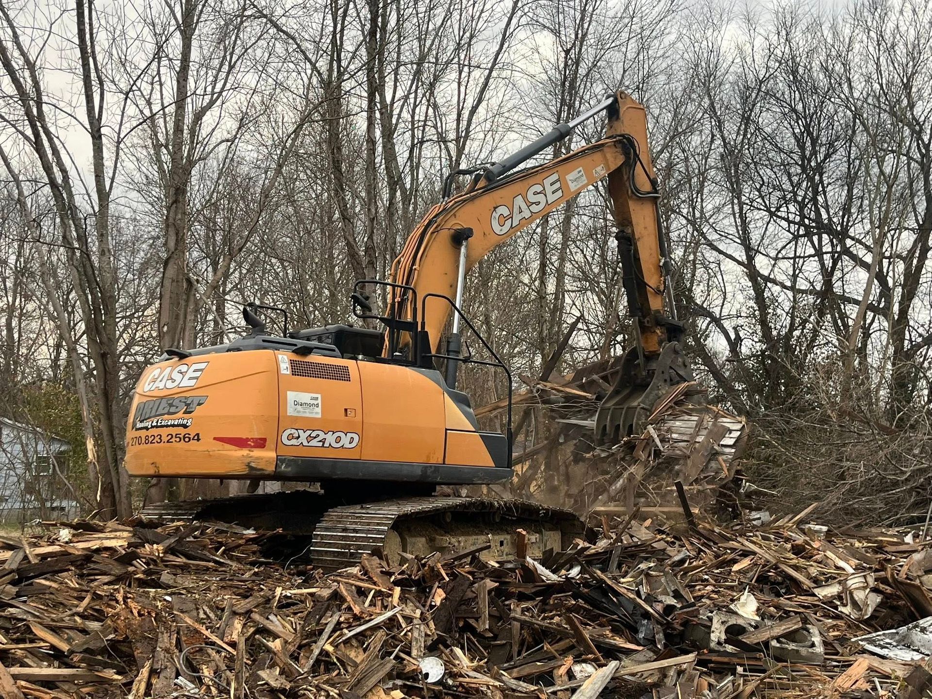 Orange excavator clearing debris in a wooded area with bare trees