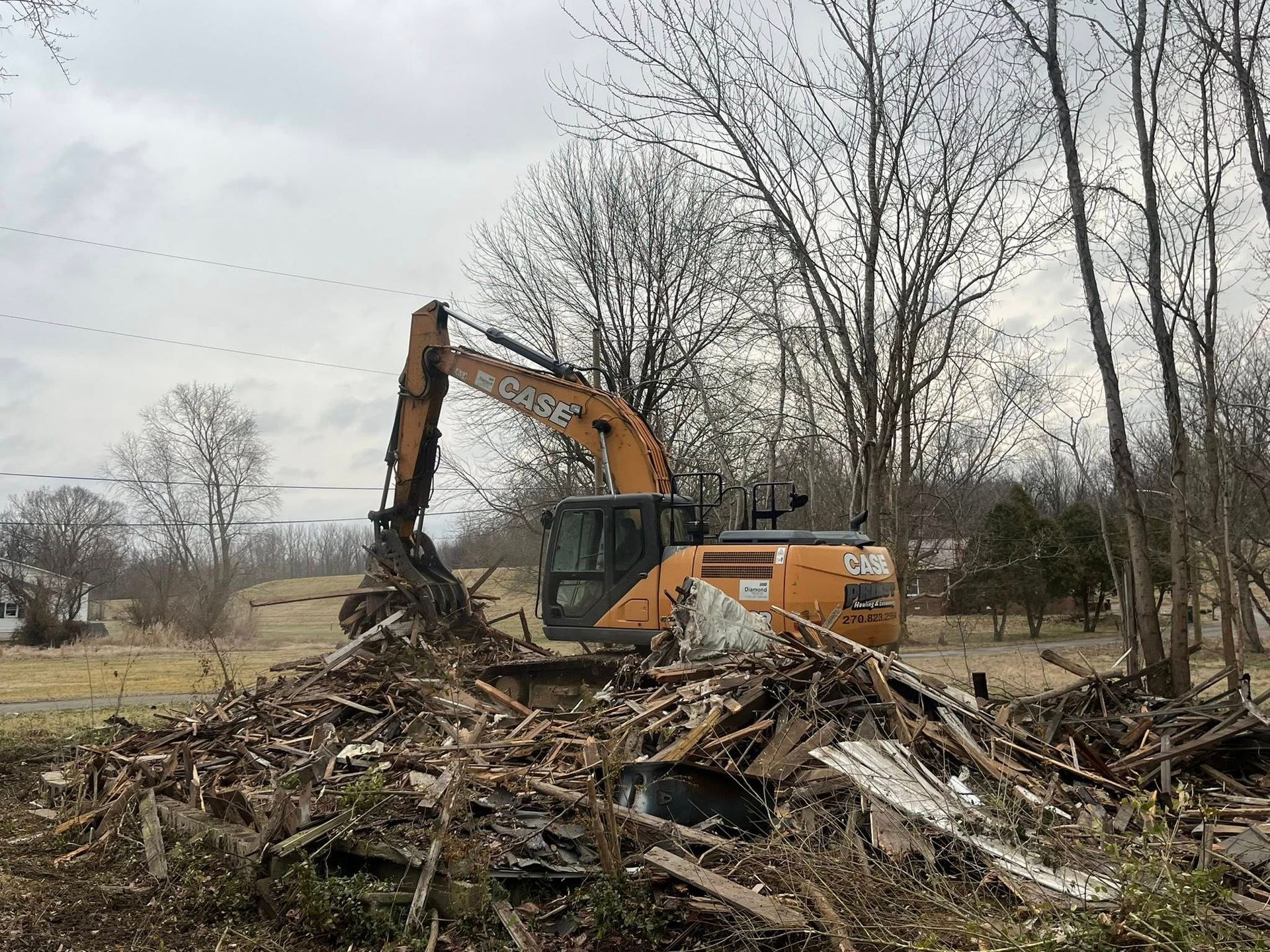 Orange excavator clearing a pile of debris in a barren lot with leafless trees behind it