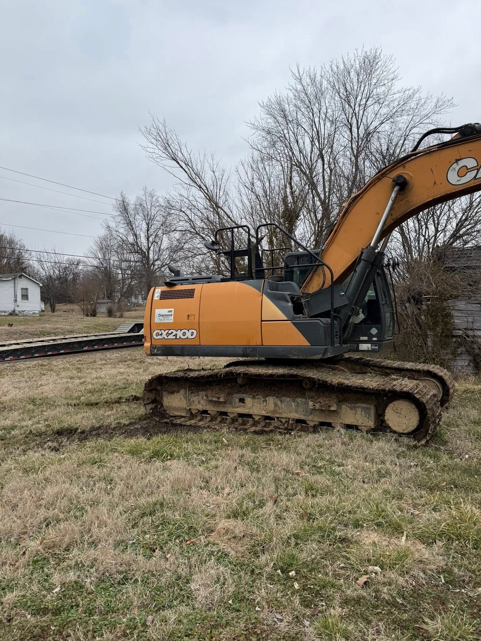 Orange excavator on tracks in a grassy field near bare trees under an overcast sky