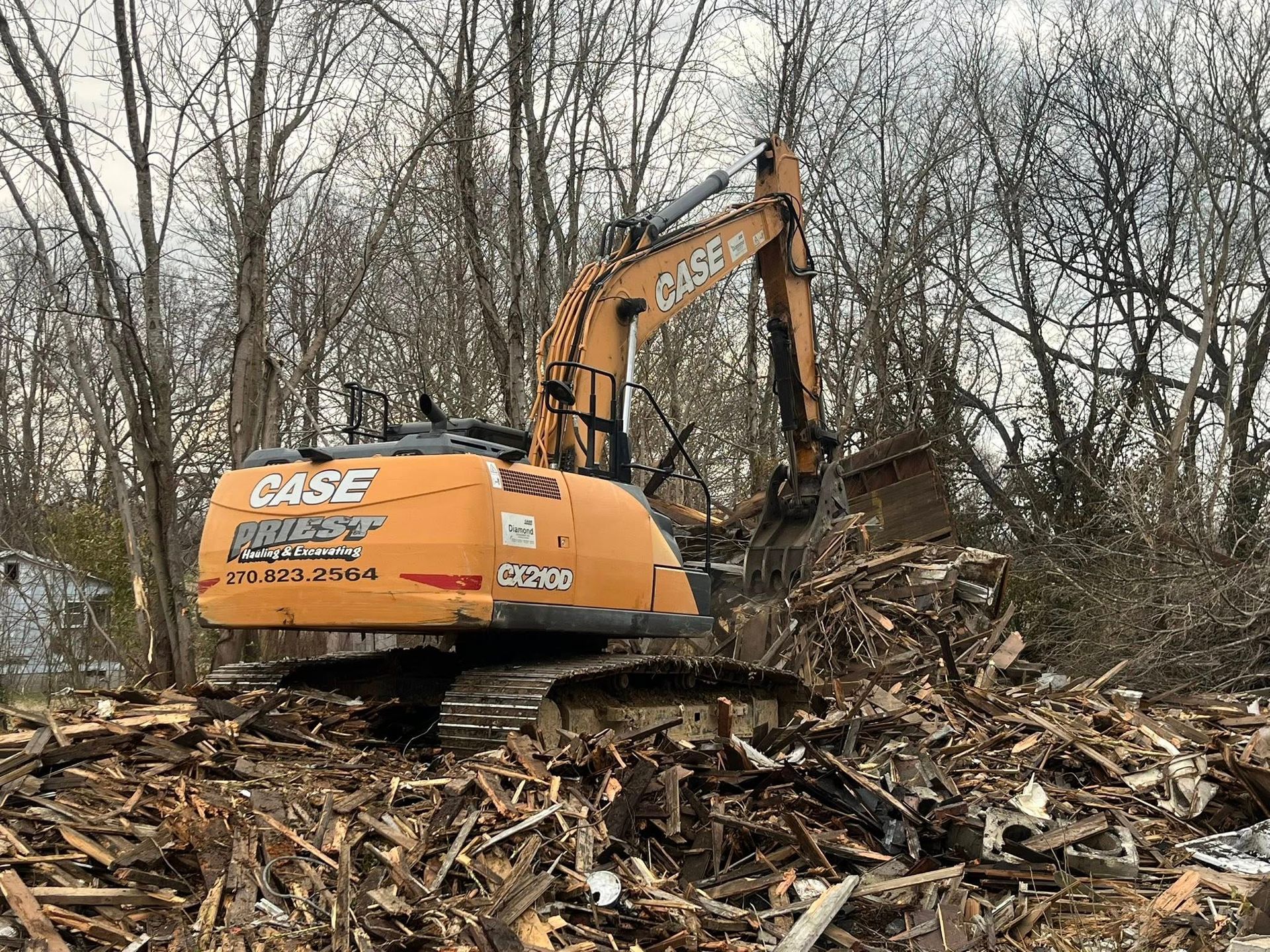 Orange excavator clearing a debris pile in a wooded area with bare trees