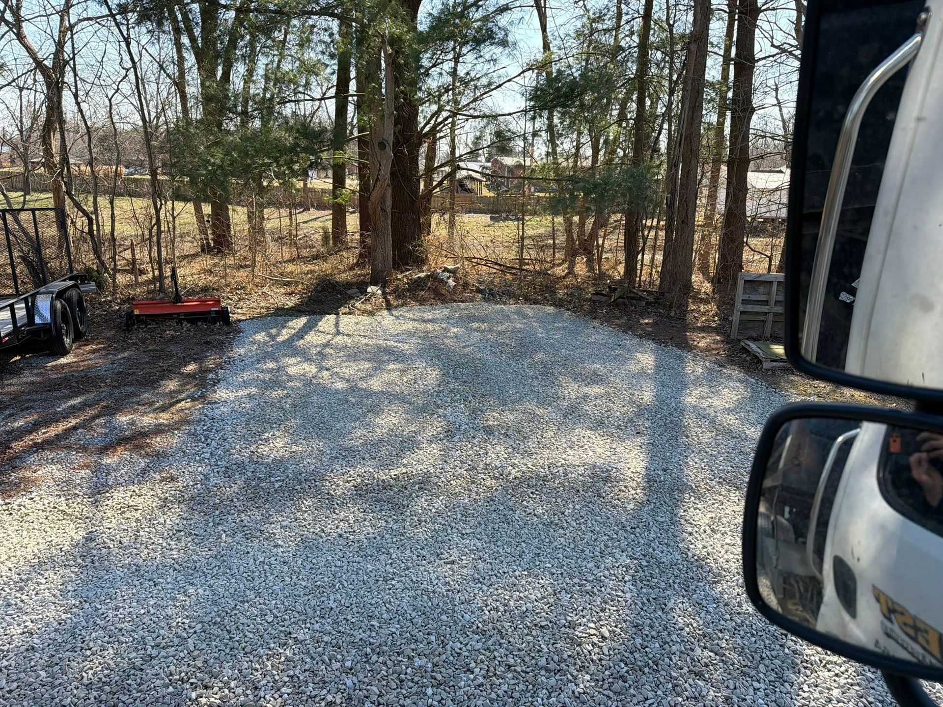 Gravel driveway beside woods, with a red cart on the left and a vehicle mirror on the right.