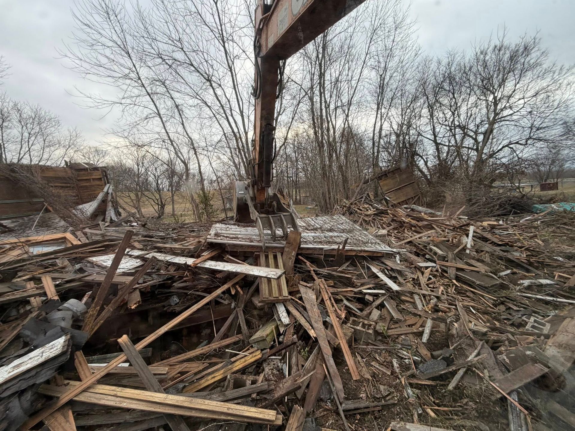 Collapsed wooden debris in a barren outdoor area under bare trees and a gray sky