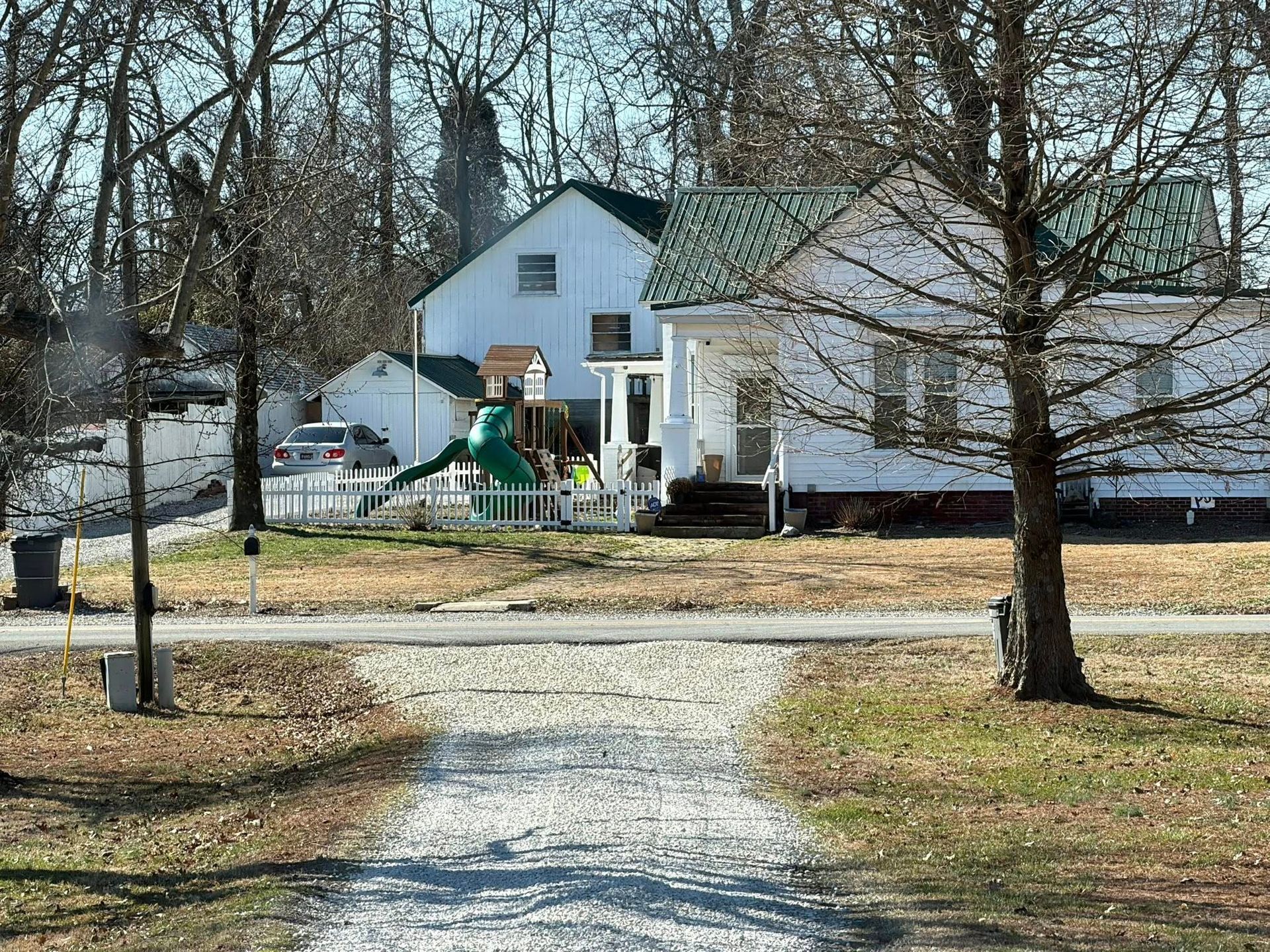 Snowy path leading to a white house with trees and a small playground in the yard