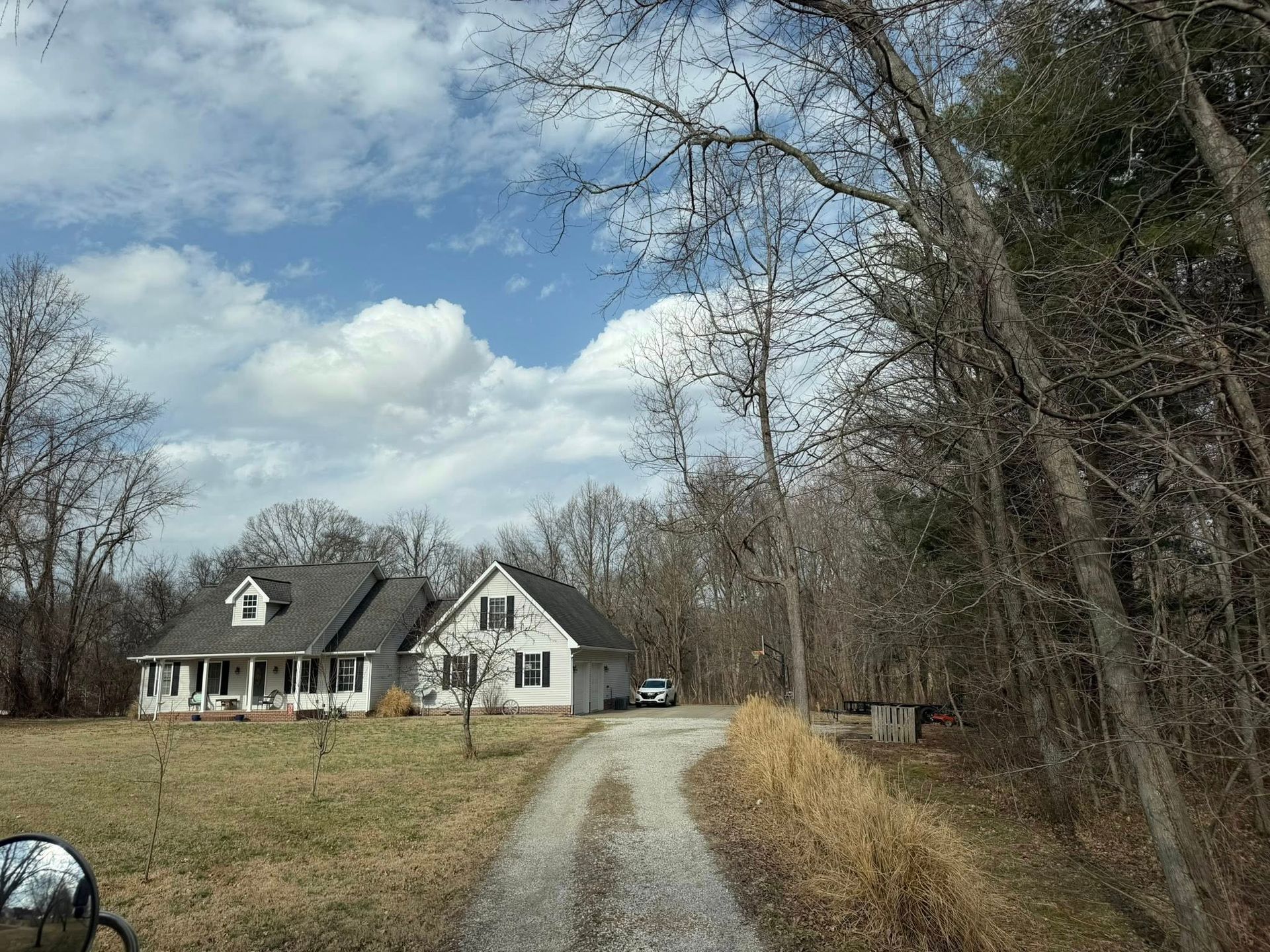 White house with a gravel driveway in a wooded rural yard under a partly cloudy sky.