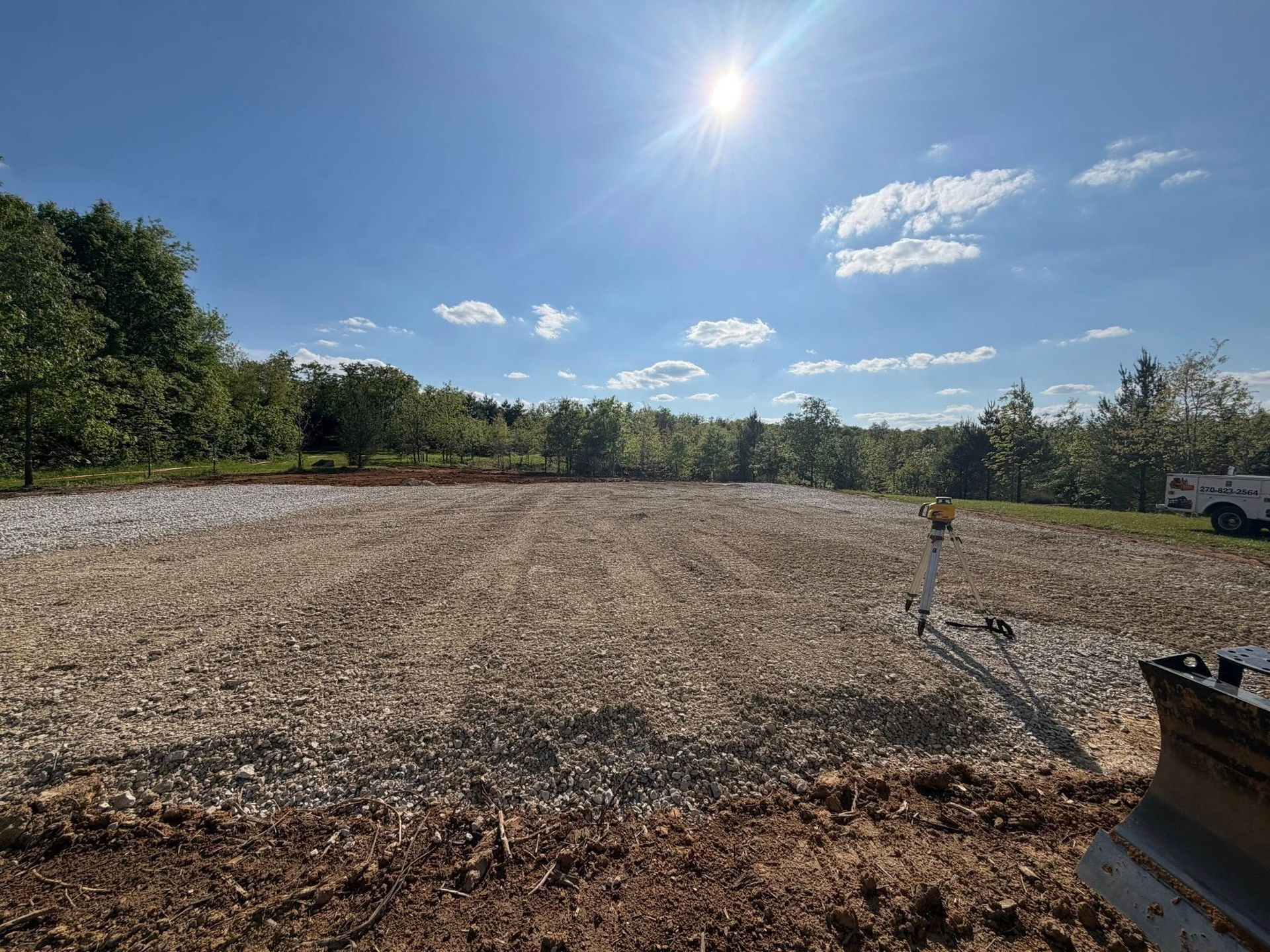 Wide gravel lot under a bright blue sky, with trees along the edges and a pipe in the foreground.